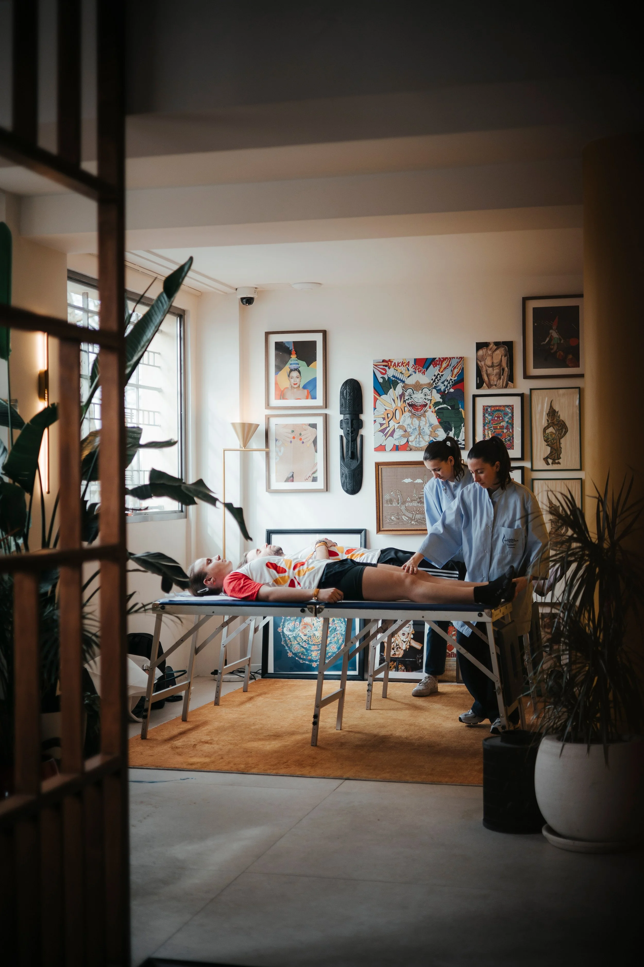 Une séance de massage ou de thérapie corporelle dans une pièce décorée d'art contemporain, avec deux praticiennes en blouse blanche travaillant sur une personne allongée sur une table.