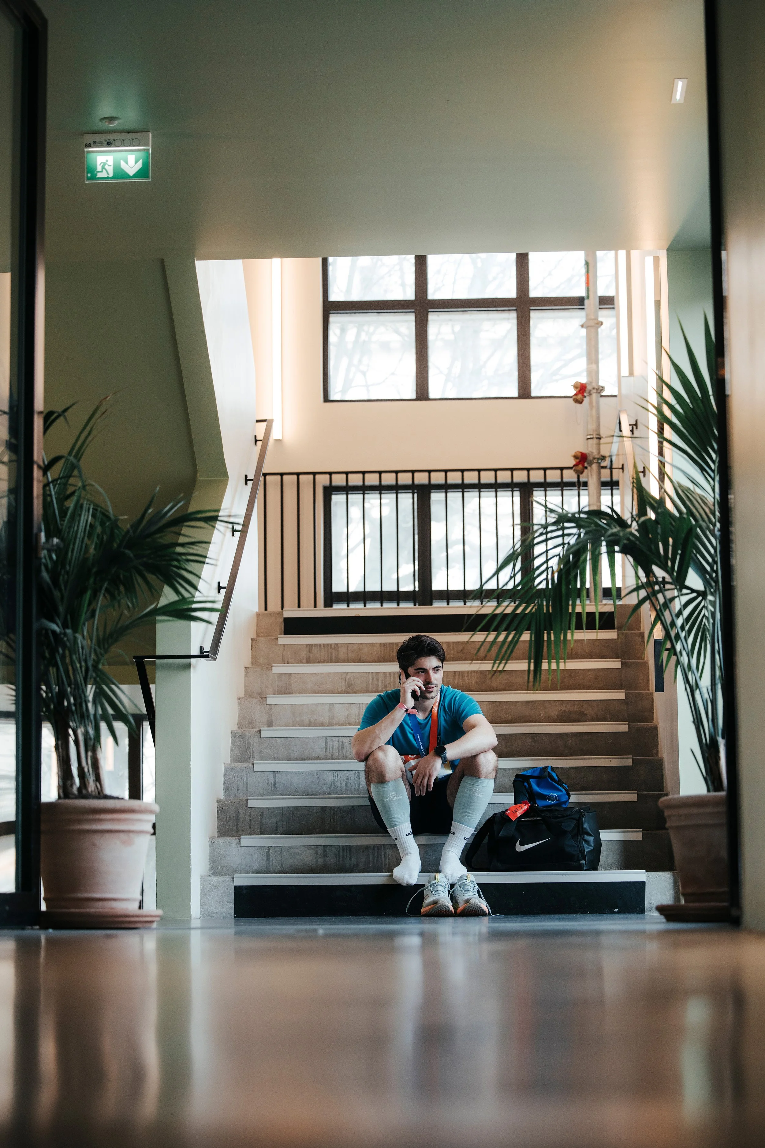 Jeune homme assis sur des escaliers d'intérieur, parlant au téléphone, portant un t-shirt bleu, des shorts noirs, des chaussettes blanches, et des chaussures de sport, avec un sac de sport noir à côté.