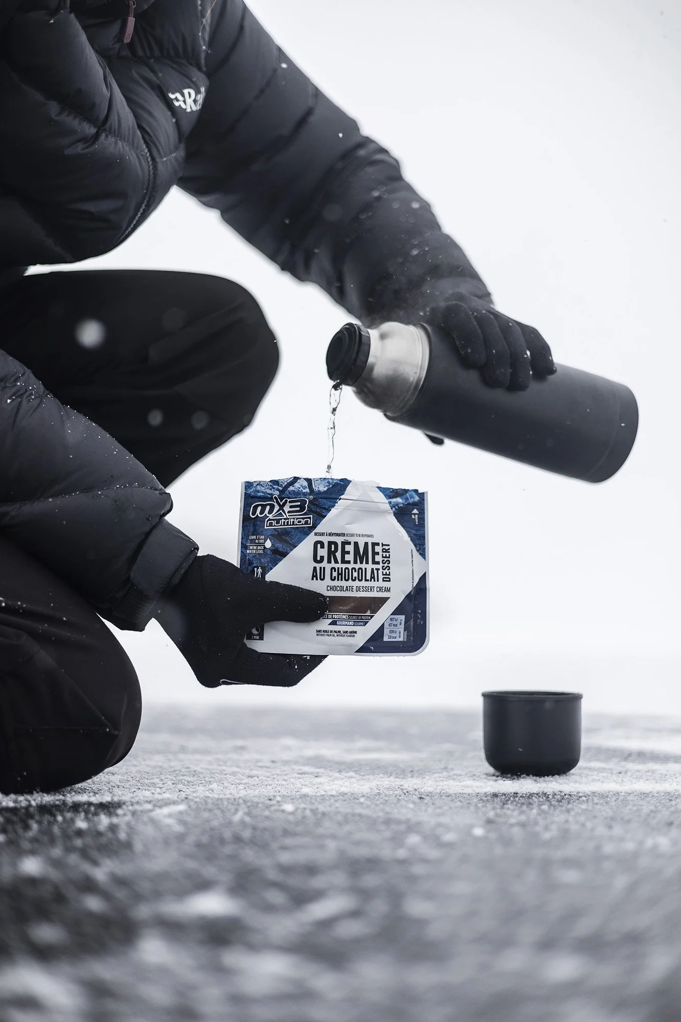 Une personne en vêtements d'hiver verse de l'eau d'une bouteille thermos dans un bol en plastique, dans un environnement enneigé.