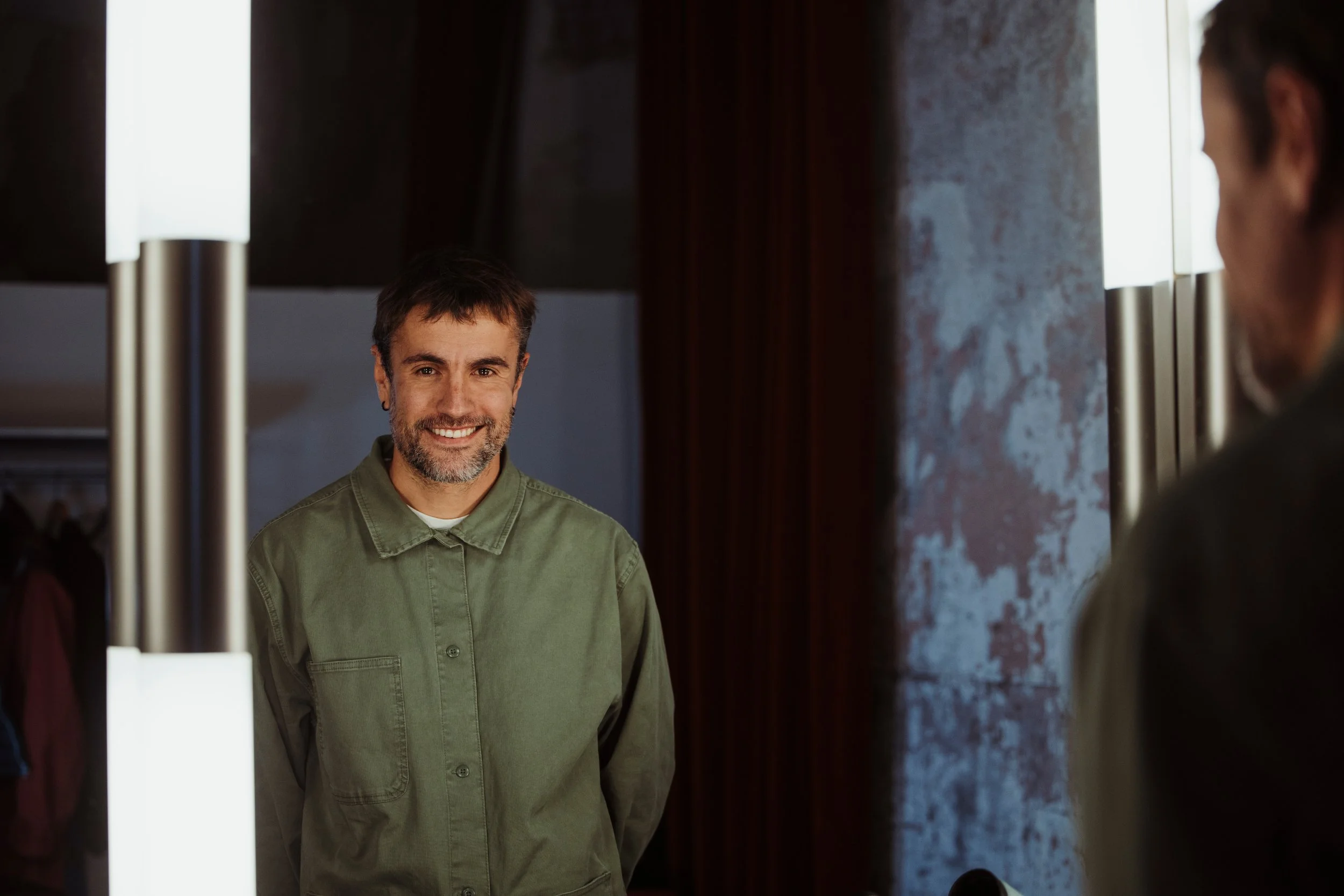 Un homme souriant se regarde dans un miroir, portant une chemise verte, dans une pièce avec un mur en texture rustique.