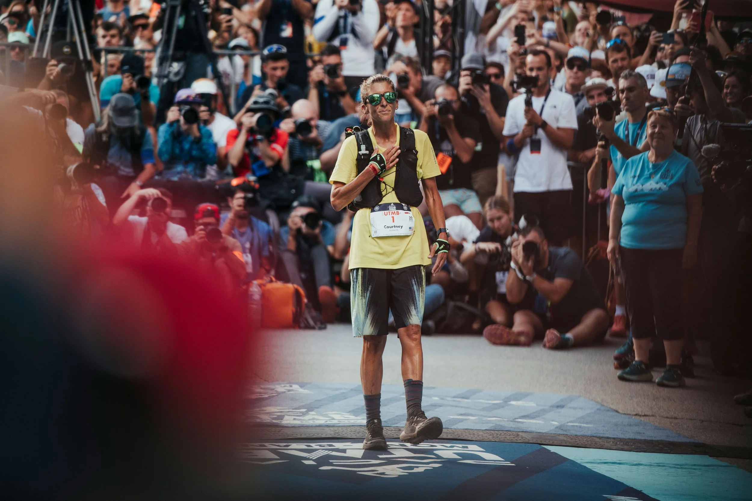 Une femme courante en tenue de sport, portant un dossard avec le nom Courtney Dauwalter, se tient sur la ligne d'arrivée en pleine couverture médiatique et entourée de nombreux journalistes et photographes.