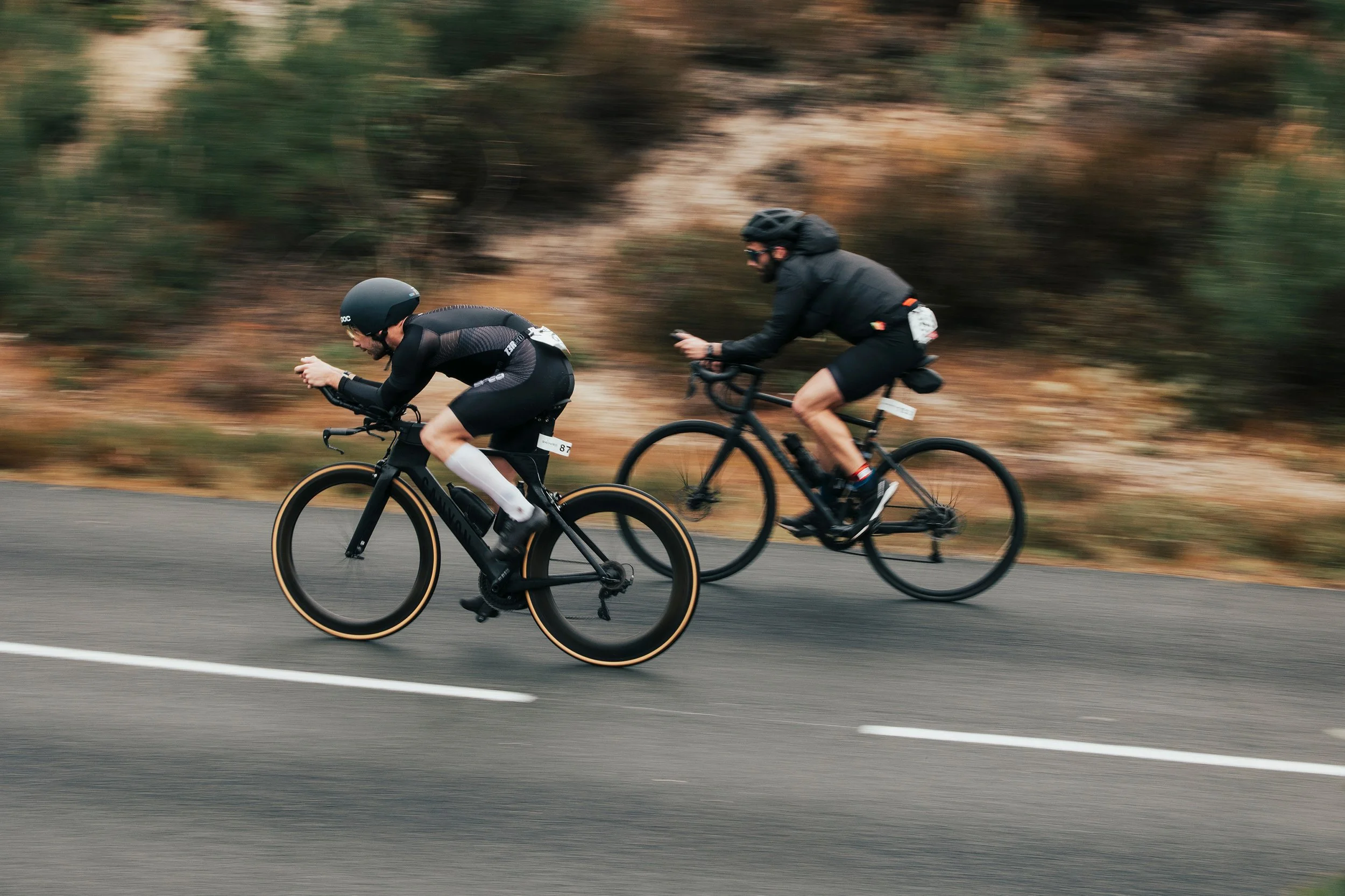 Deux cyclistes en compétition sur une route entièrement goudronnée entourée d'une végétation de saison automnale.
