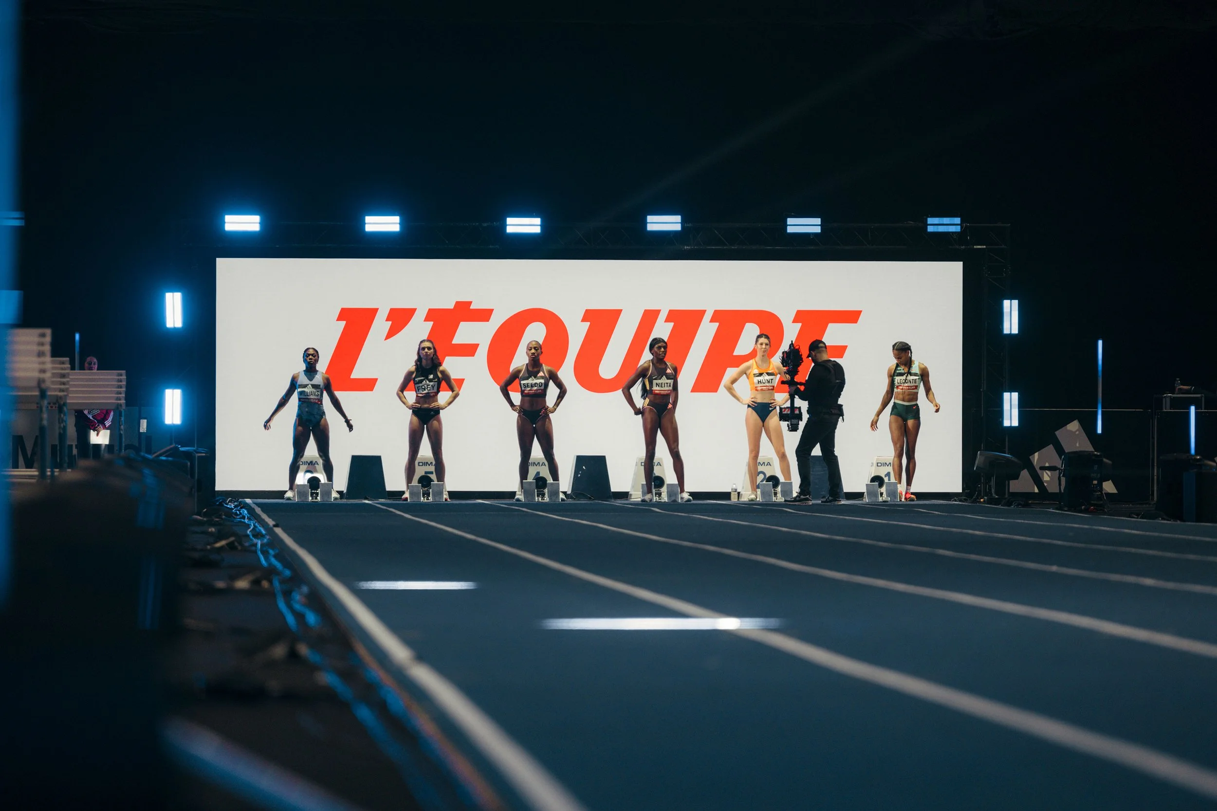 Coureuses sur la ligne de départ d'une compétition d'athlétisme, avec un panneau lumineux affichant 'L'ÉQUIPE' en arrière-plan.