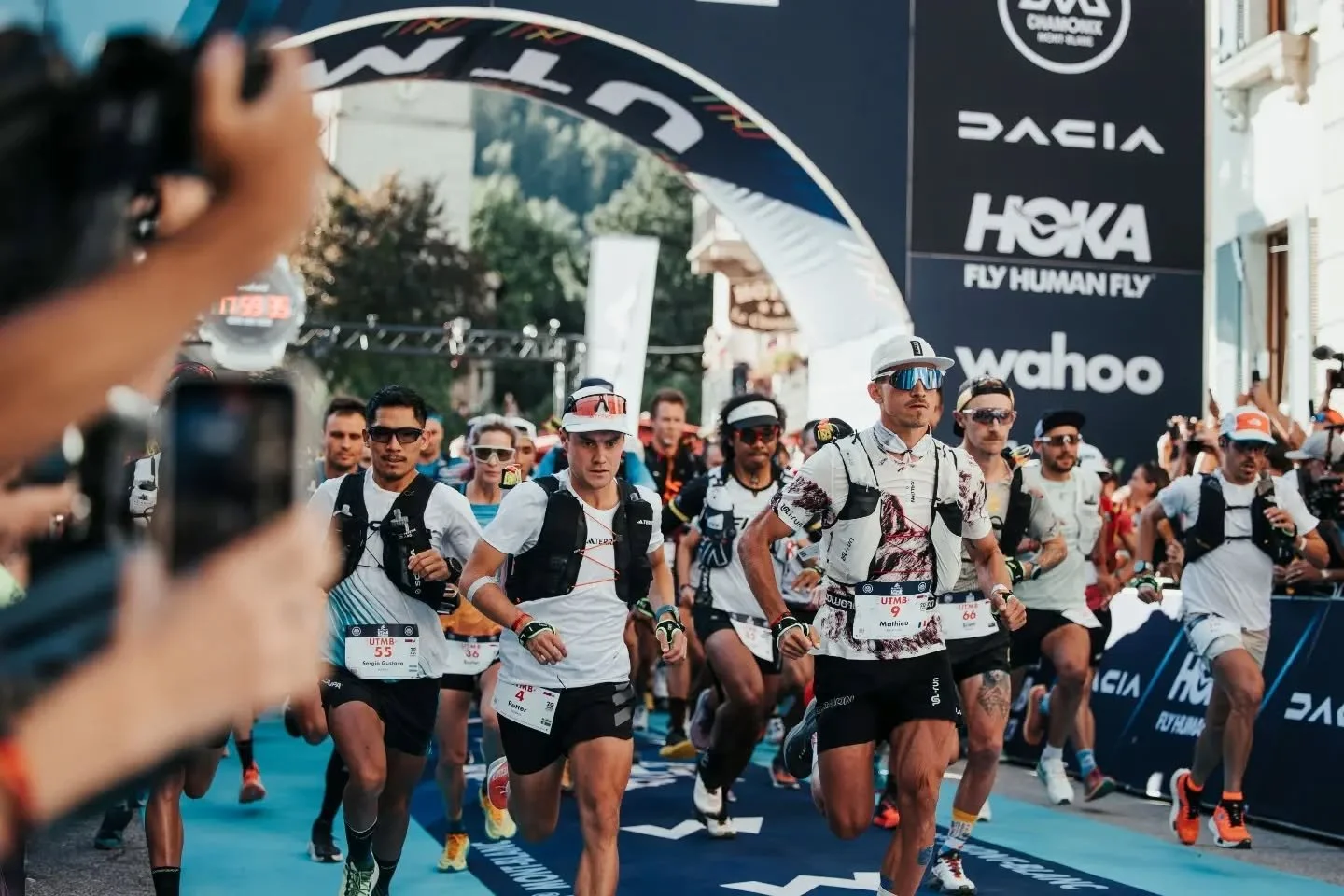 Des coureurs participant à un marathon ou une course, courant sous une arche de départ ou d'arrivée. Certains portent des bibs avec des numéros. L'événement semble très animé avec un public en arrière-plan.