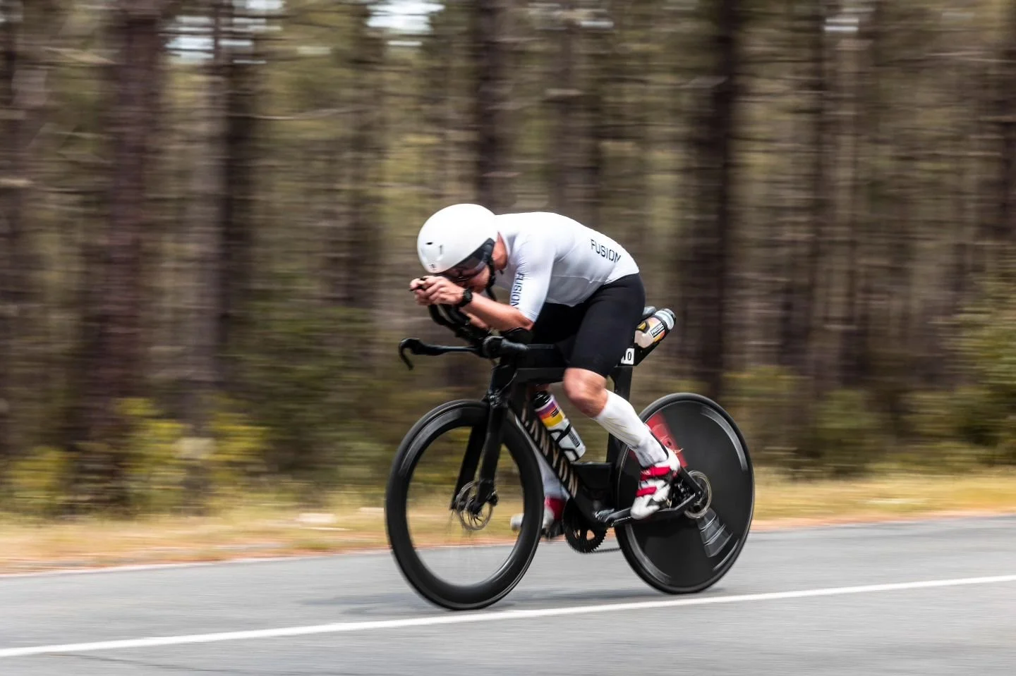 Un cycliste sur un vélo de course en pleine vitesse sur une route, portant un casque blanc et un maillot blanc et noir, avec un arrière-plan flou représentant une forêt.