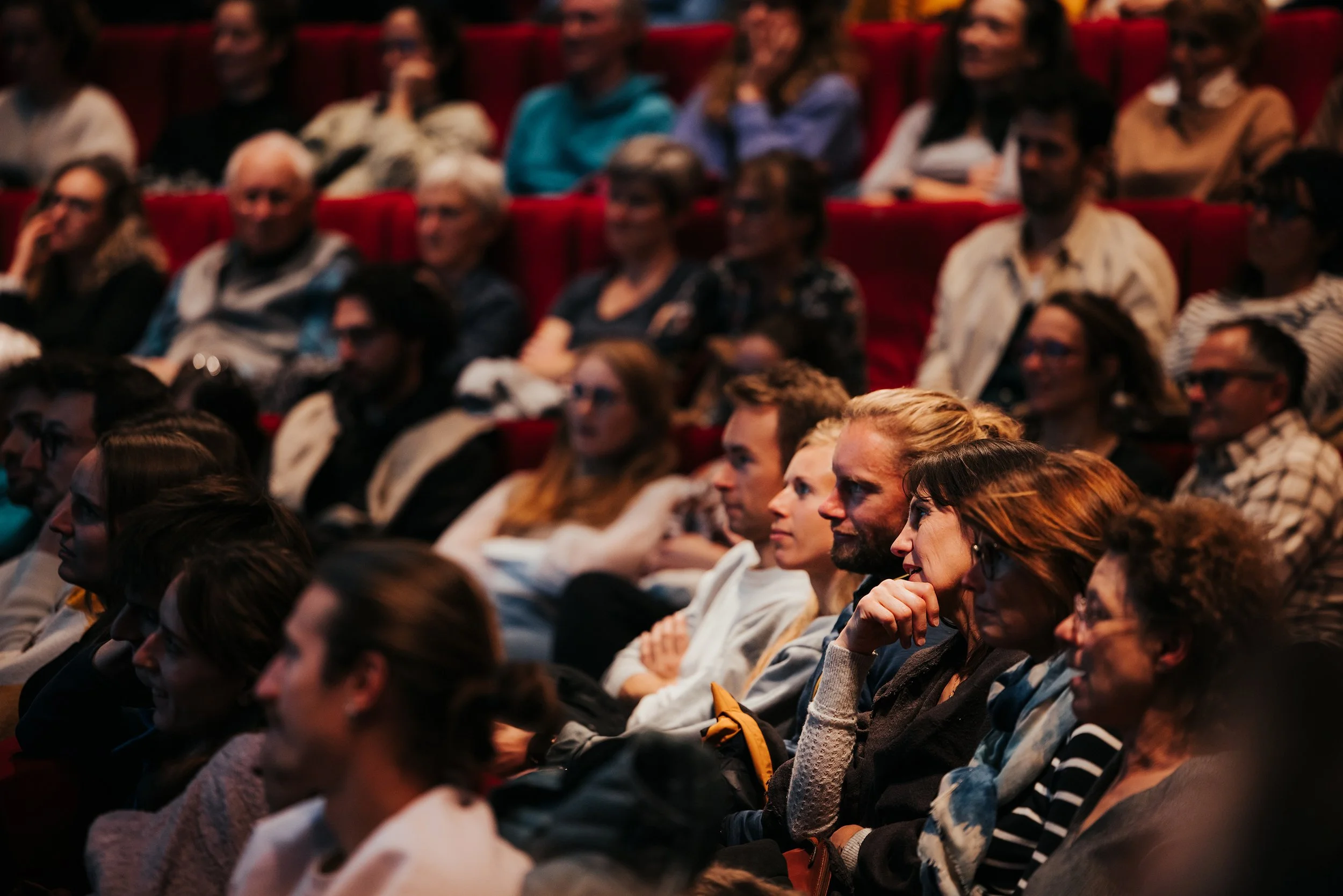Public attentif dans un auditorium sombre avec des sièges rouges, regardant un spectacle ou une conférence.