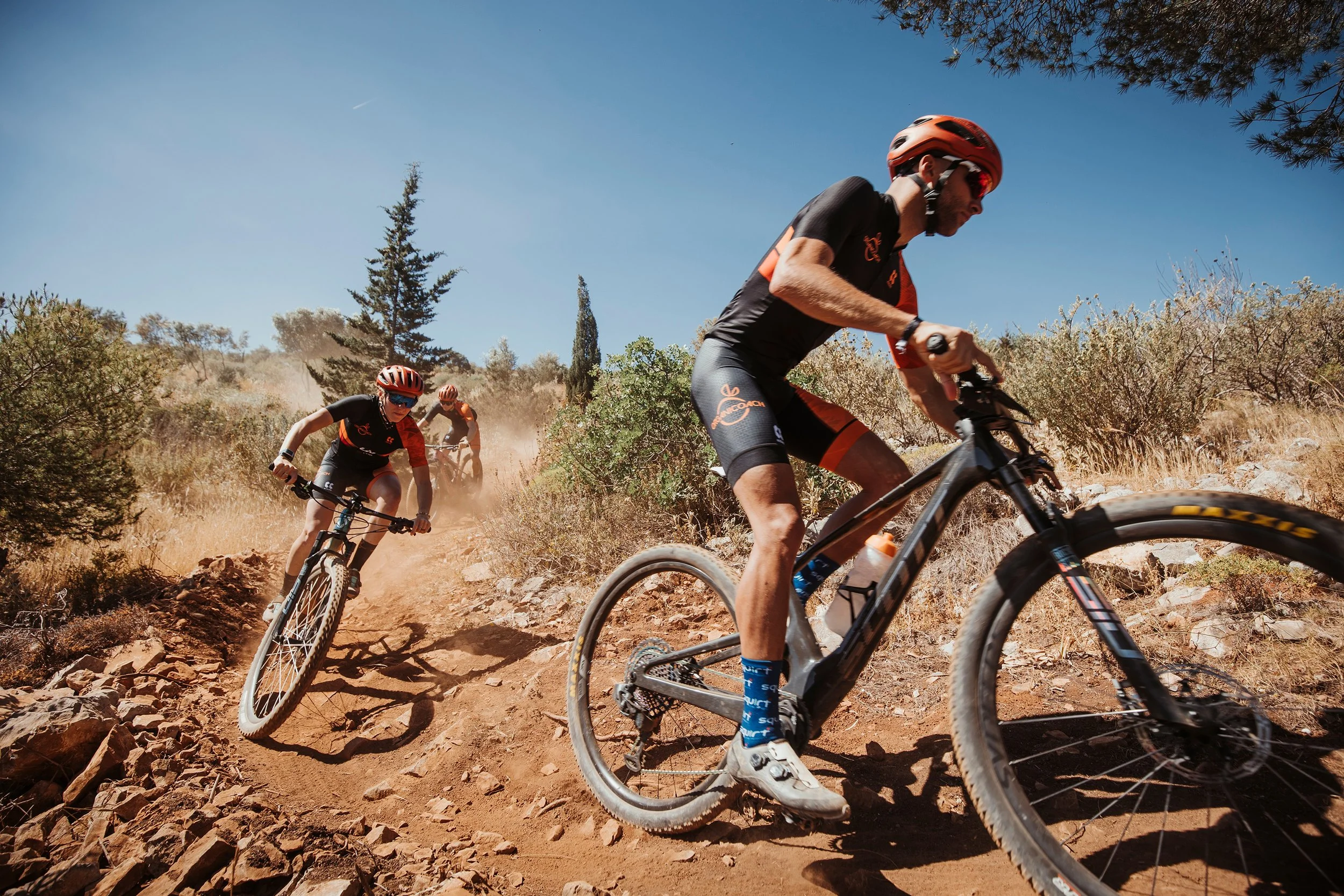 Trois cyclistes en train de descendre une pente rocheuse en montagne, portant des casques et des vêtements de sport, avec un ciel bleu et des arbustes secs en arrière-plan.