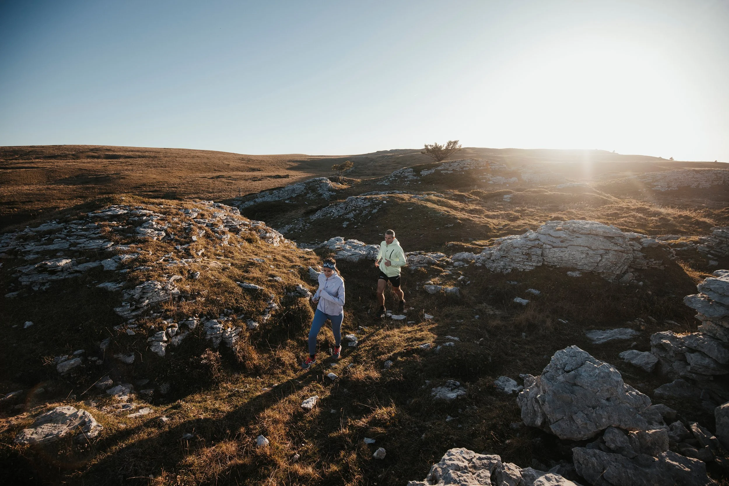 Deux personnes courent sur un sentier de montagne au lever ou coucher du soleil, avec des rochers et des collines en arrière-plan.