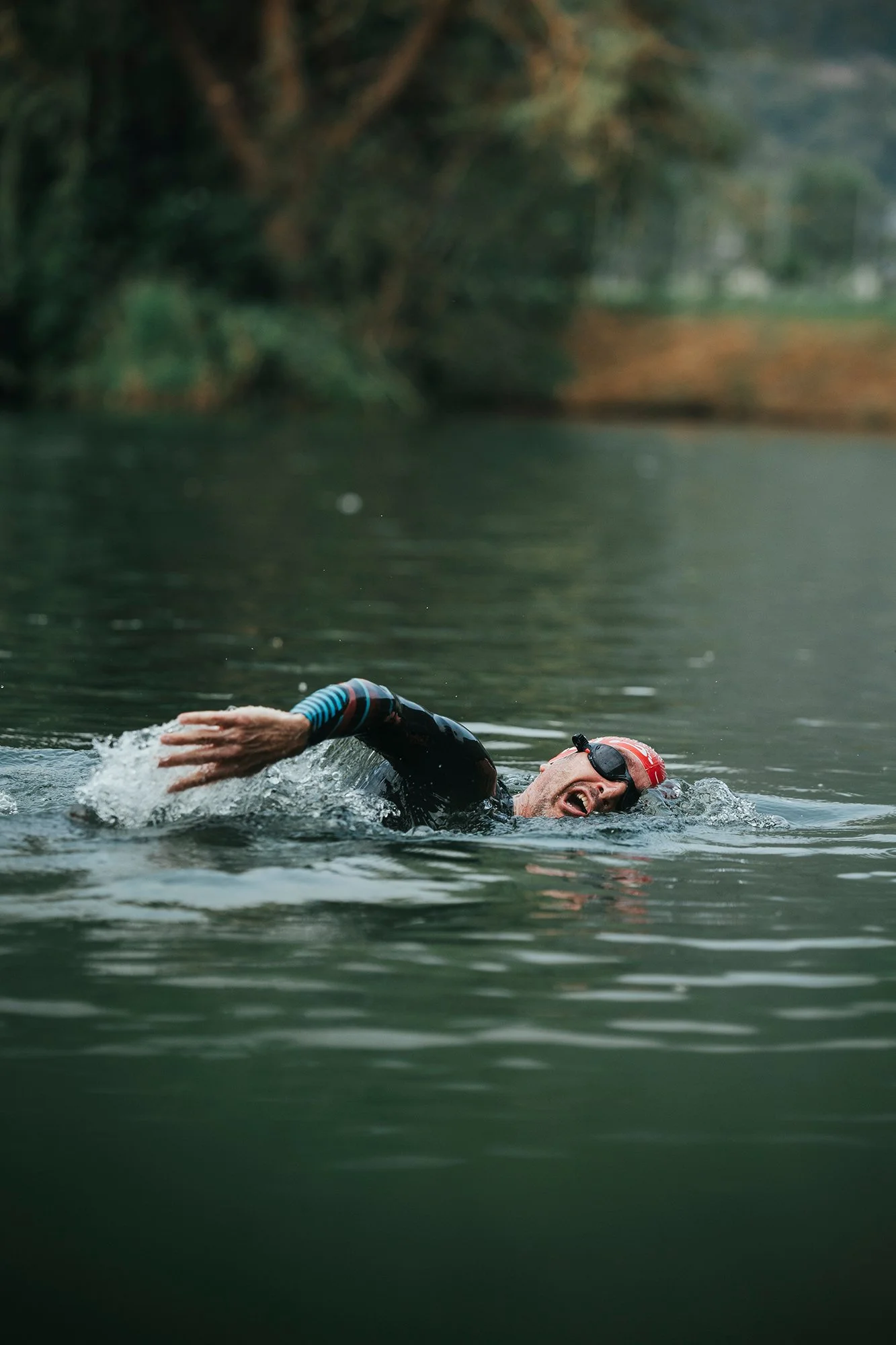 Un homme nageant dans une étendue d'eau, portant une combinaison de natation, lunettes de natation, et un bonnet rouge, en pleine activité de natation, avec un environnement naturel en arrière-plan.