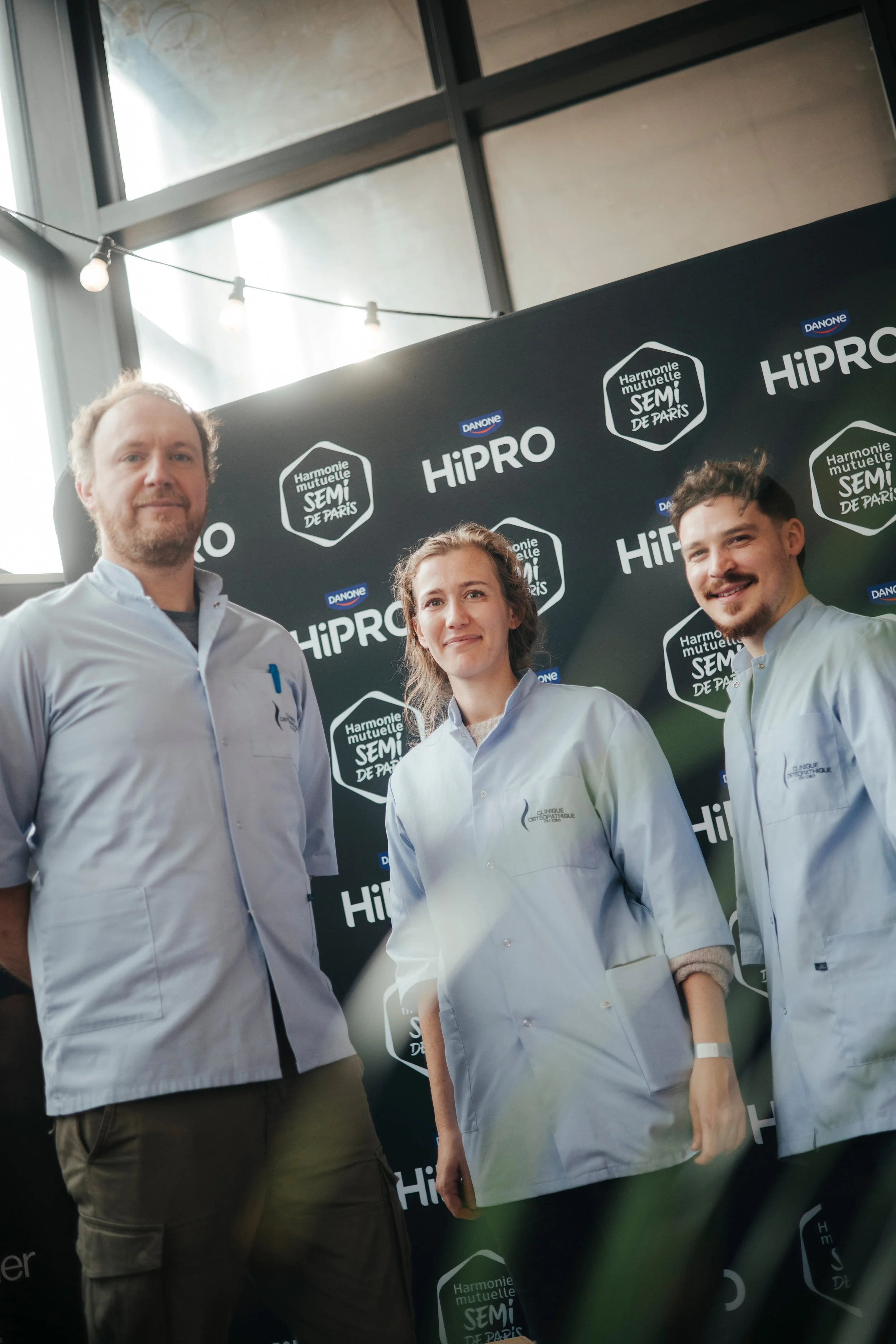 Trois personnes portant des blouses blanches posent devant un fond noir avec des logos de Danone, HiPRO et une harmonie mutuelle semi de Paris, dans un environnement intérieur avec de grandes fenêtres et un éclairage suspendu.