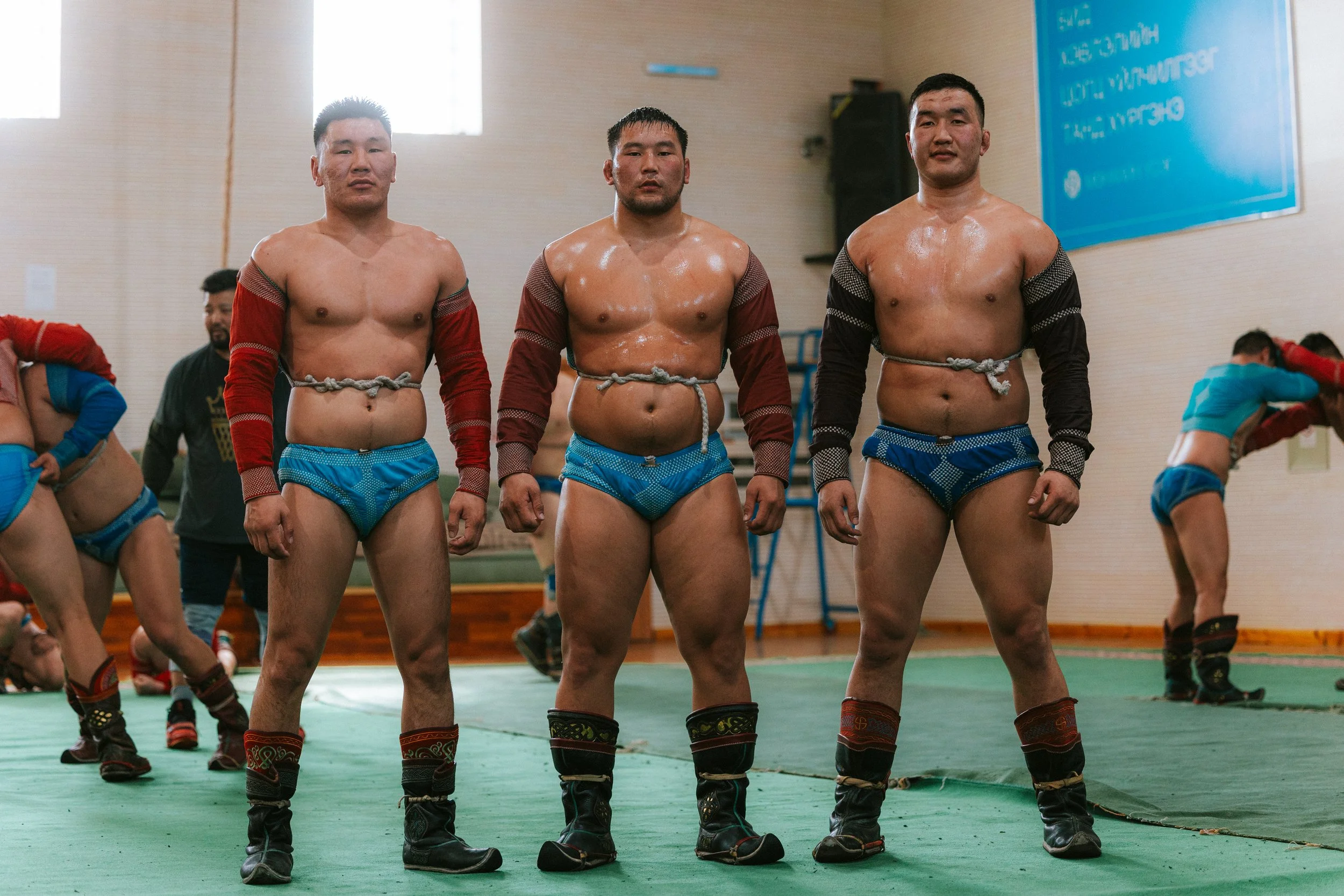 Trois hommes mongols, nus de la taille à la tête, portant des bottes traditionnelles et des vêtements de sport, se tiennent en rang dans un gymnase. Ils ont l'air fatigué mais concentré.