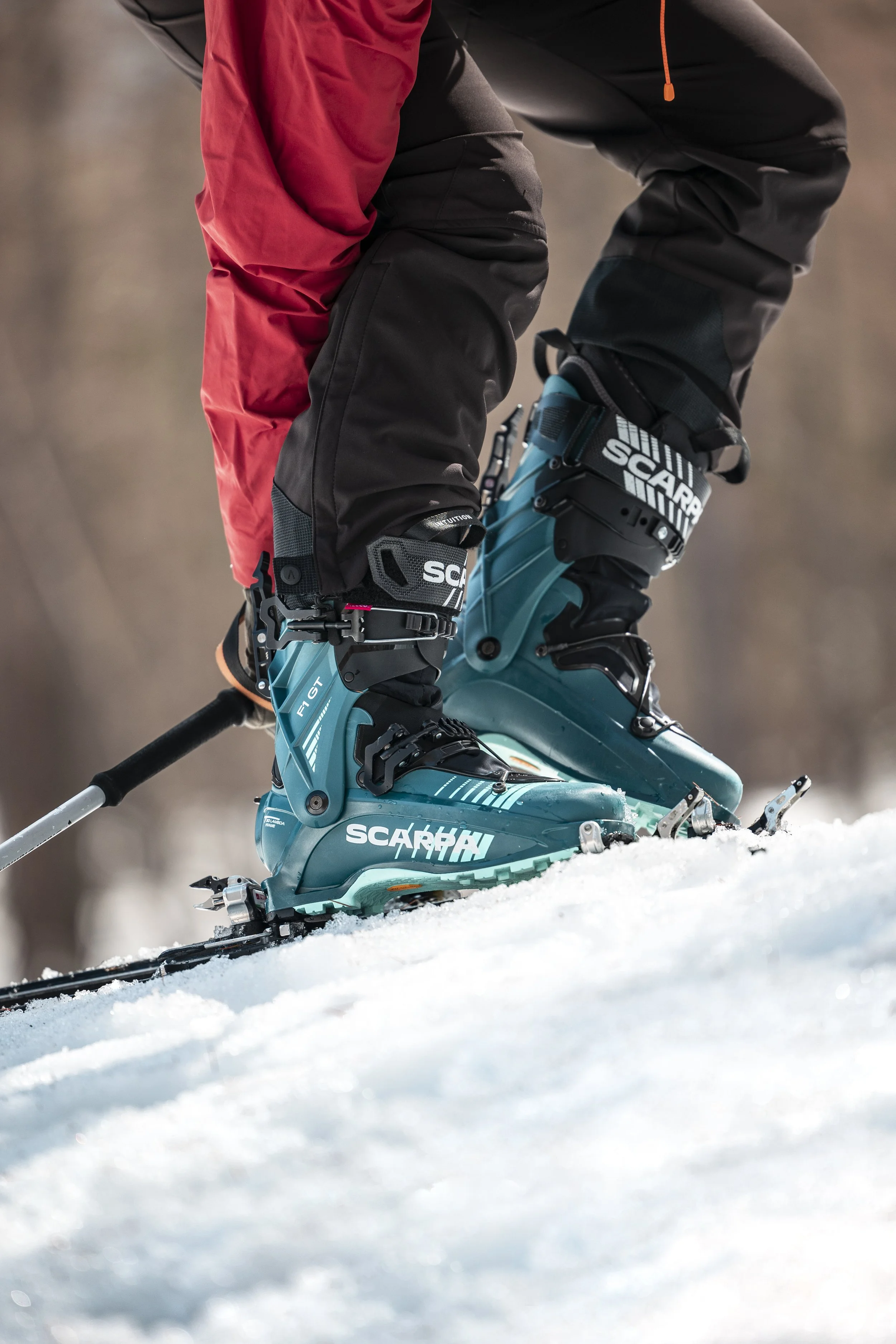 Chaussures de ski alpins de marque Scarpa, portées par une personne dans la neige, avec des pantalons de ski noirs et un vêtement rouge.