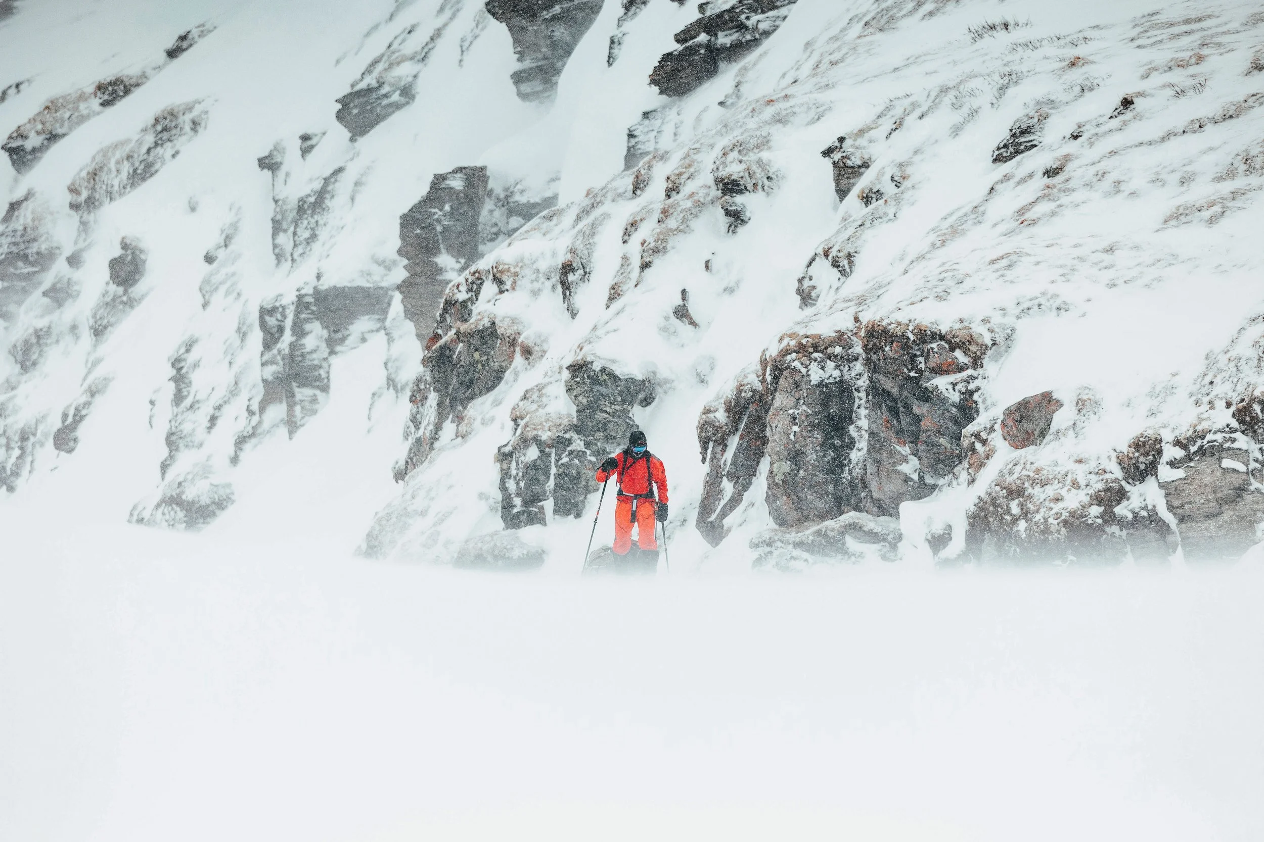 Un skieur en tenue orange avec des skis, dans un paysage enneigé avec des rochers et une falaise rocheuse.
