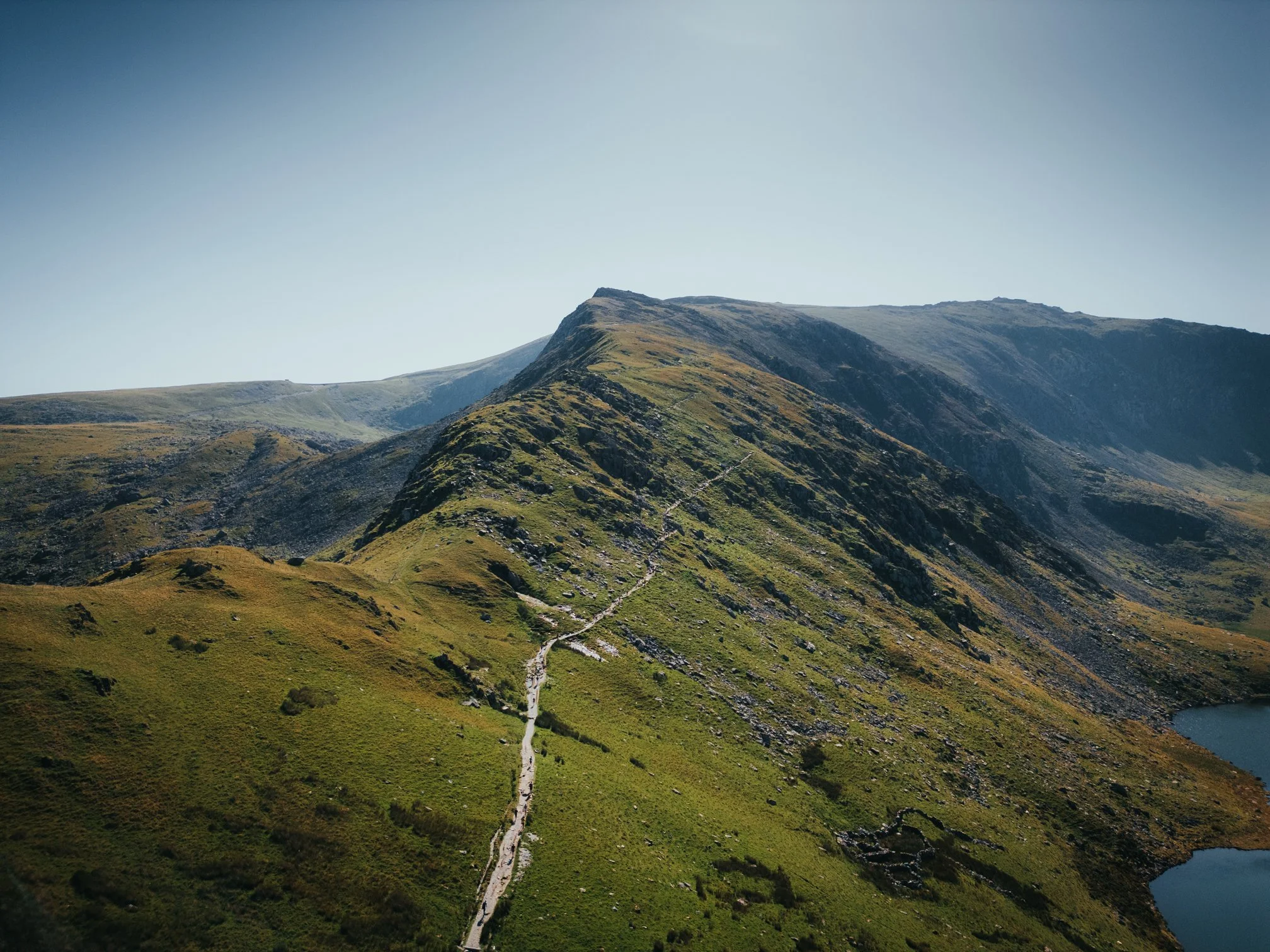 Une montagne surplombant une vallée avec un sentier serpentant vers le sommet et un lac en contrebas.