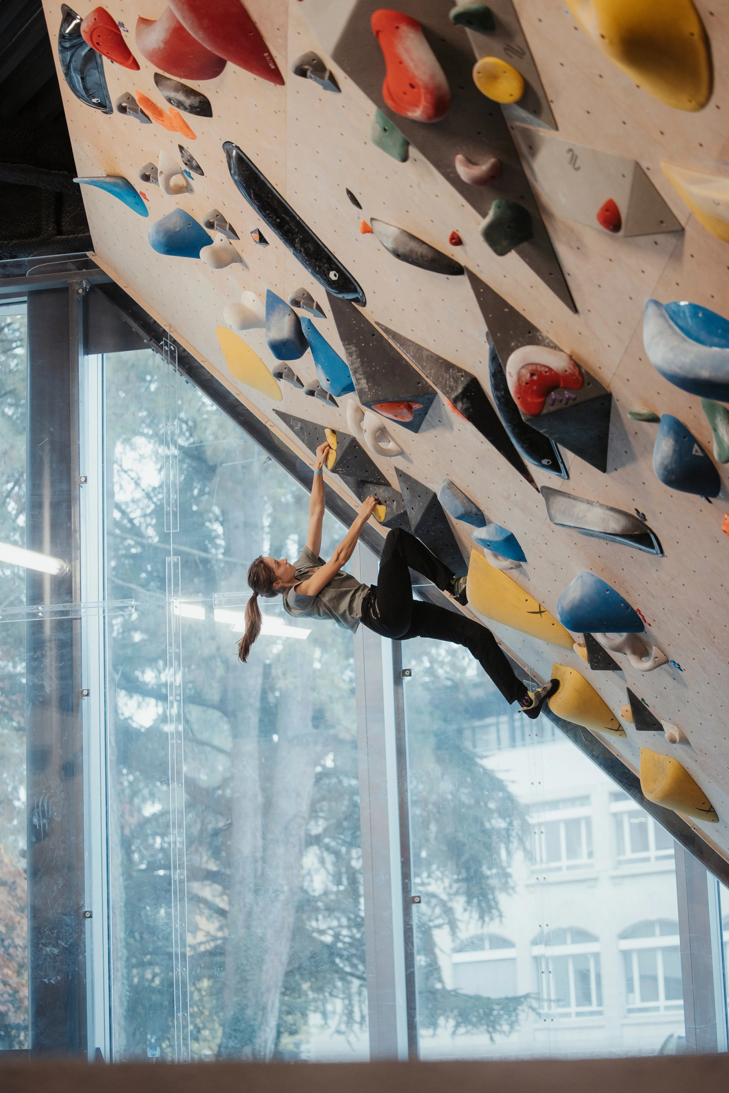 Une femme grimpe une paroi d'escalade intérieure, avec plusieurs prises colorées sur un mur en bois, près d'une grande fenêtre laissant entrer la lumière naturelle. Salle ATOM à Annecy.