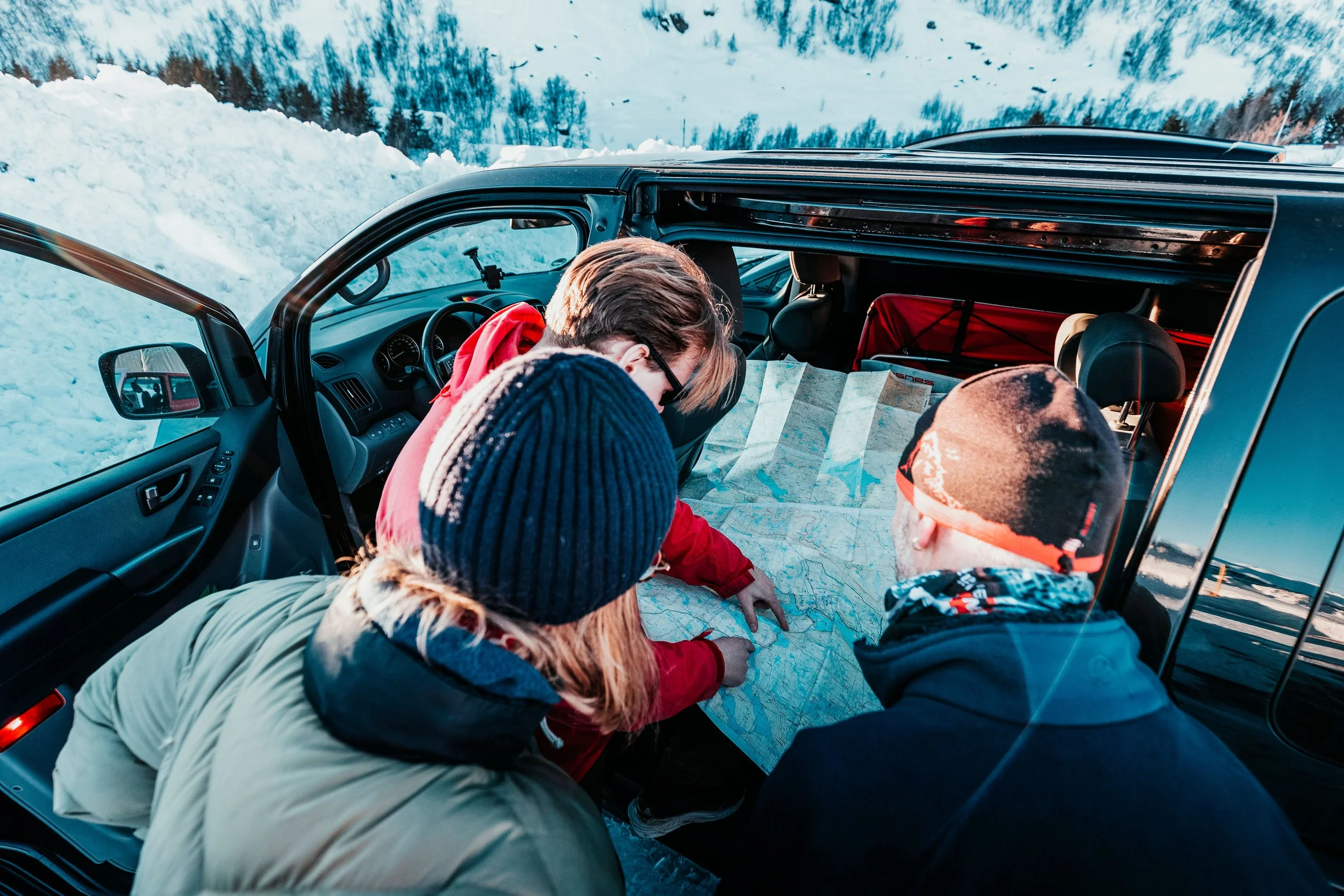 Quatre personnes en vêtements d'hiver regardent une carte routière dans une voiture stationnée dans un paysage enneigé avec des montagnes.