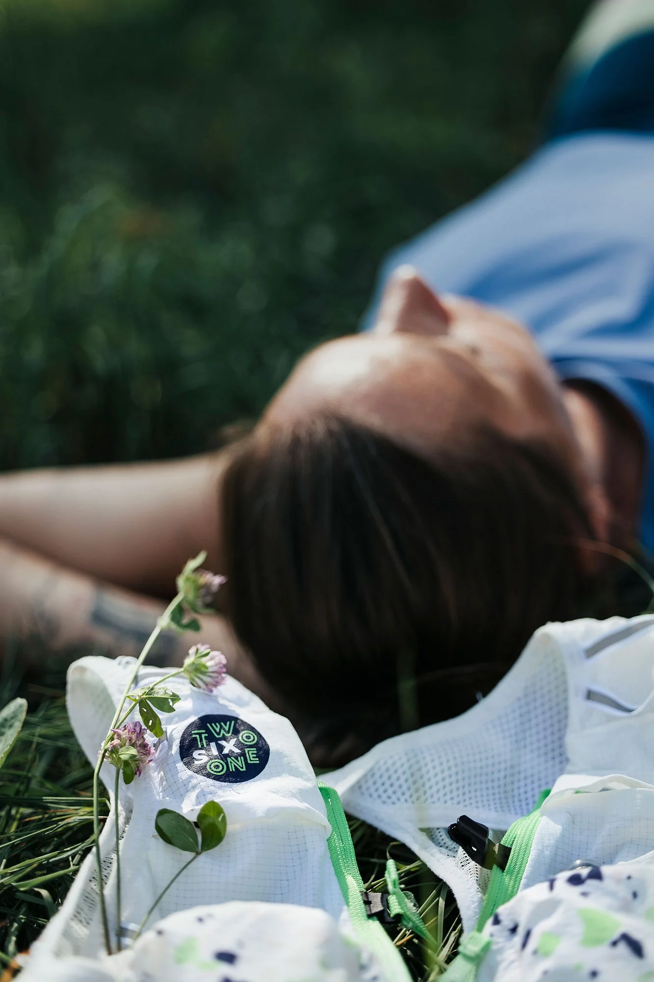 Une personne allongée sur l'herbe avec des vêtements de sport autour, dont un chiffon blanc avec un logo "Two Six One" et une petite branche de fleurs violettes.
