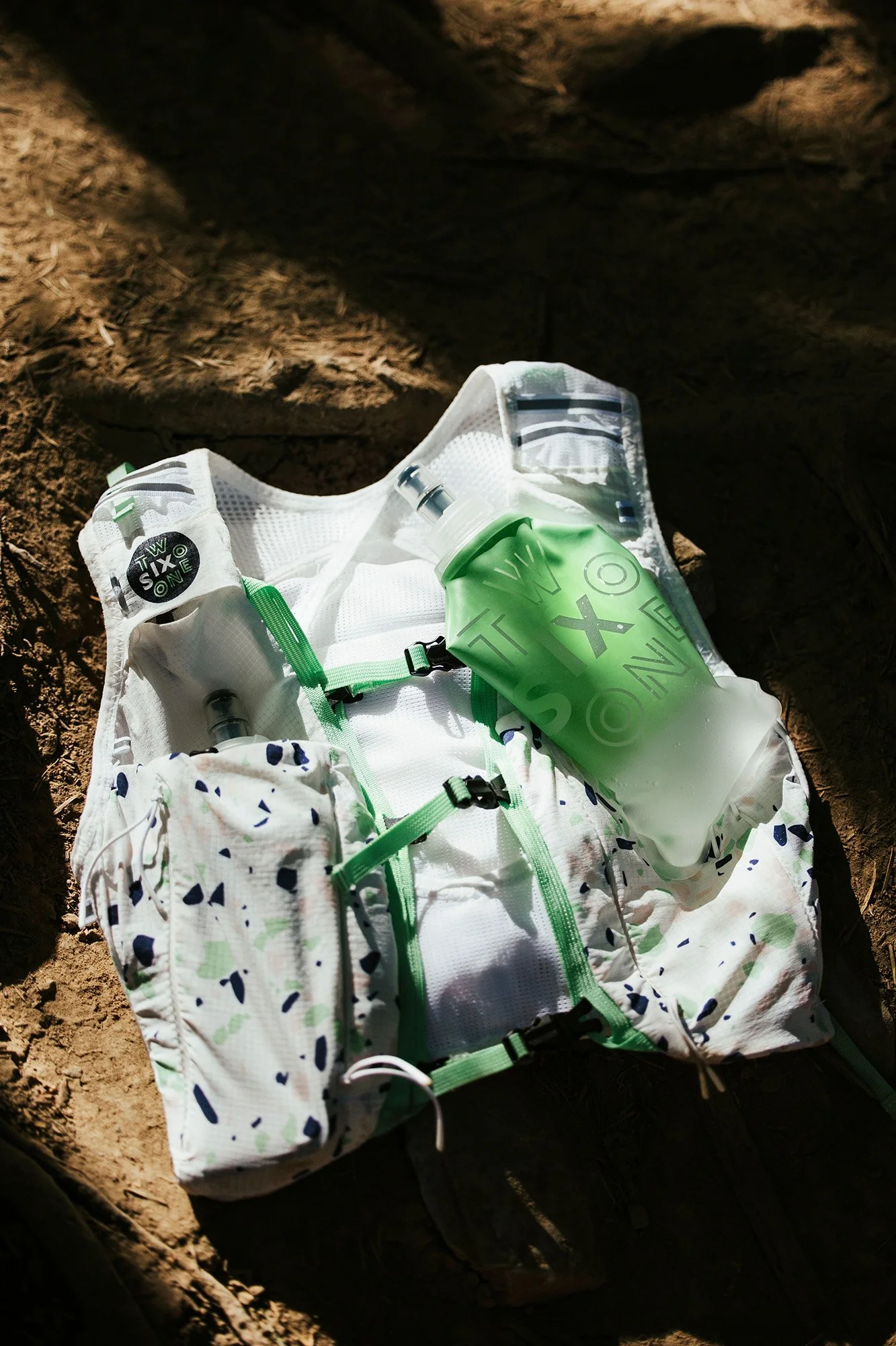 Un sac à dos blanc avec des accents verts, contenant une gourde verte, une fiole et d'autres petits objets, posé sur le sol en terre dans un endroit ombragé.