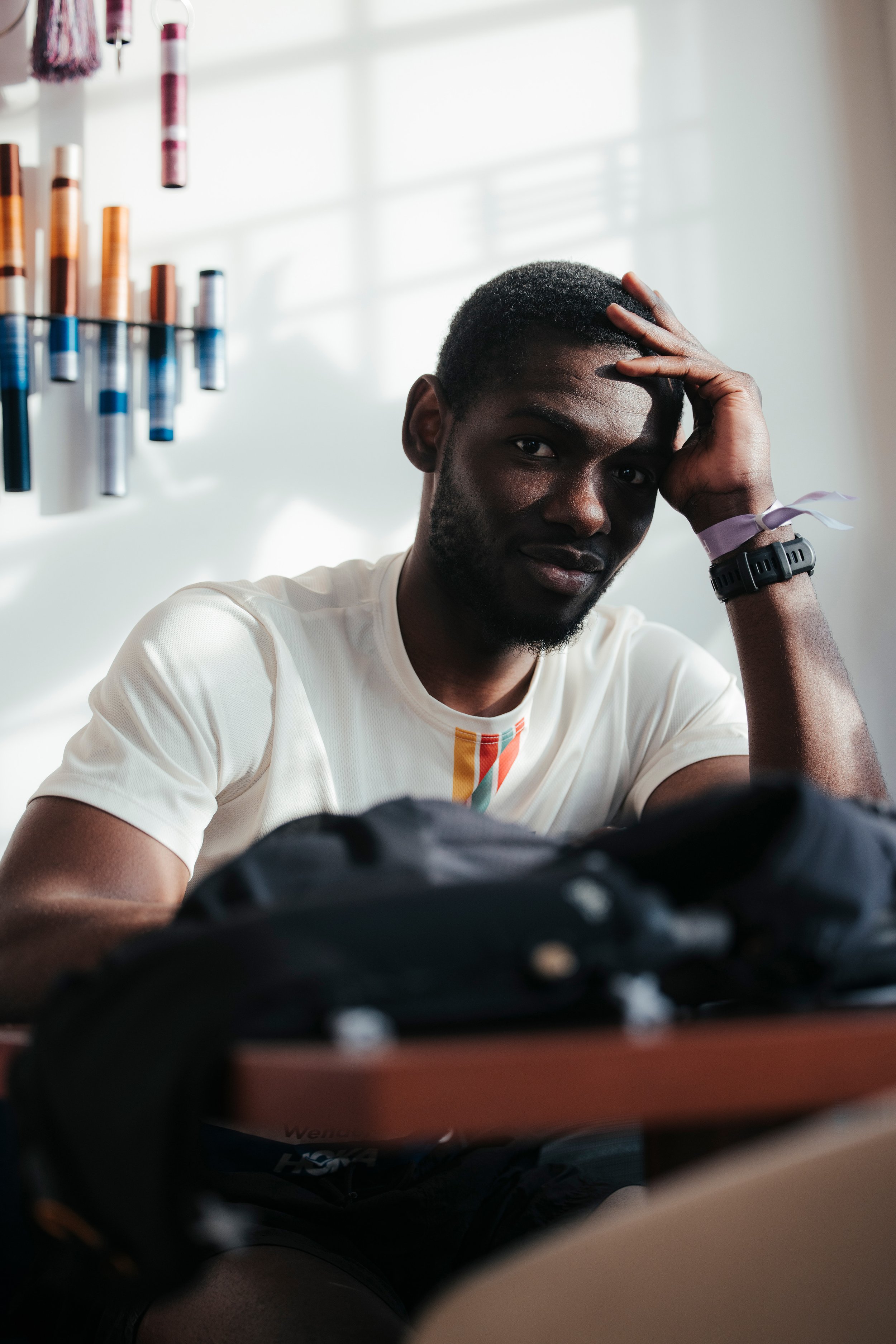 Un homme, un coureur, assis à une table, avec une main sur la tête, dans une pièce lumineuse avec des rouleaux de peinture suspendus au mur derrière lui.