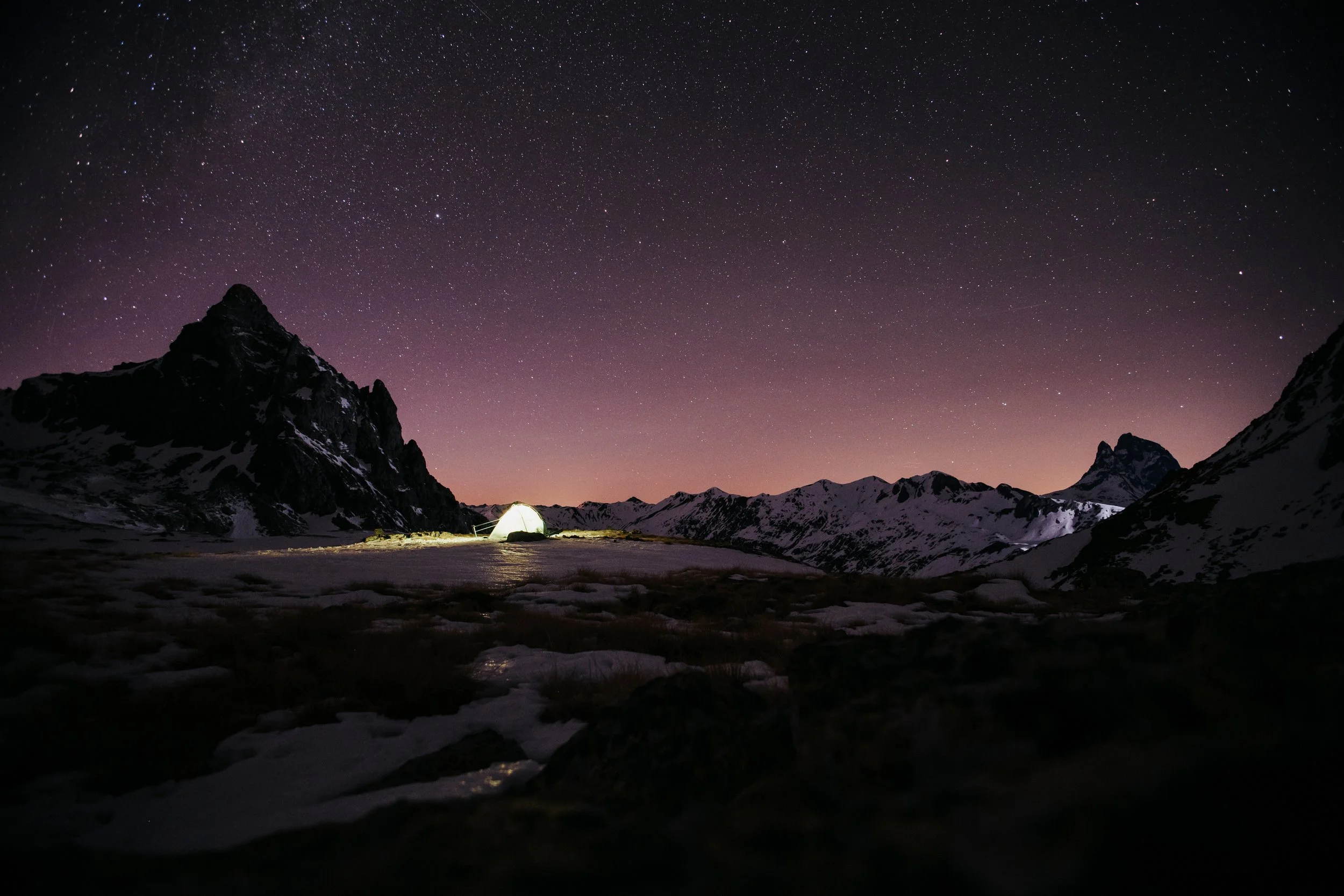Tente éclairée dans un paysage de montagnes enneigées sous un ciel étoilé au crépuscule.