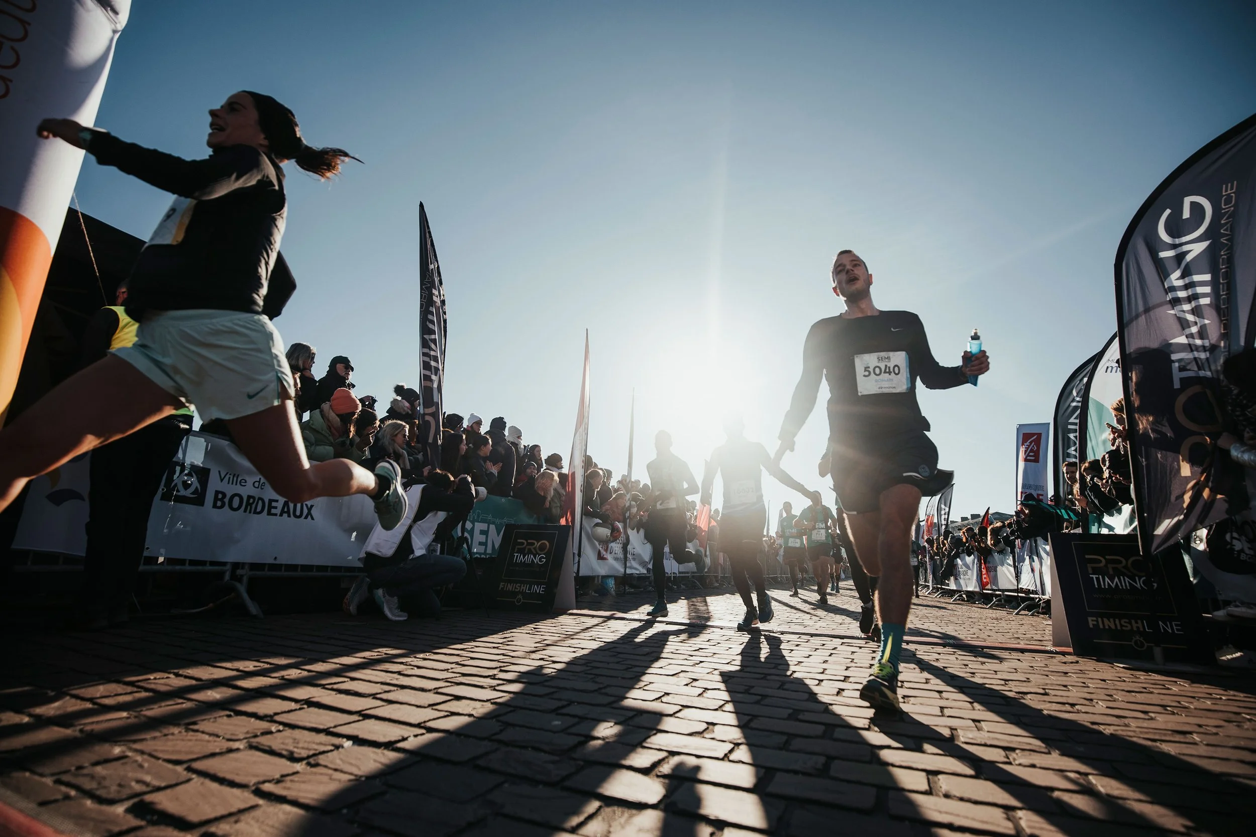 Des coureurs franchissent la ligne d'arrivée lors d'une course, avec des spectateurs applaudissant et des drapeaux en arrière-plan, sous un ciel ensoleillé.