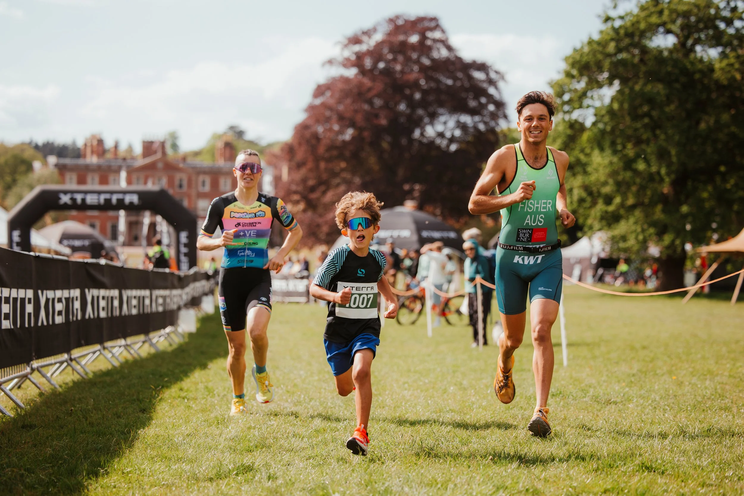 Trois coureurs en pleine course lors d'un événement sportif en plein air, avec un arbre au centre et un bâtiment en arrière-plan.