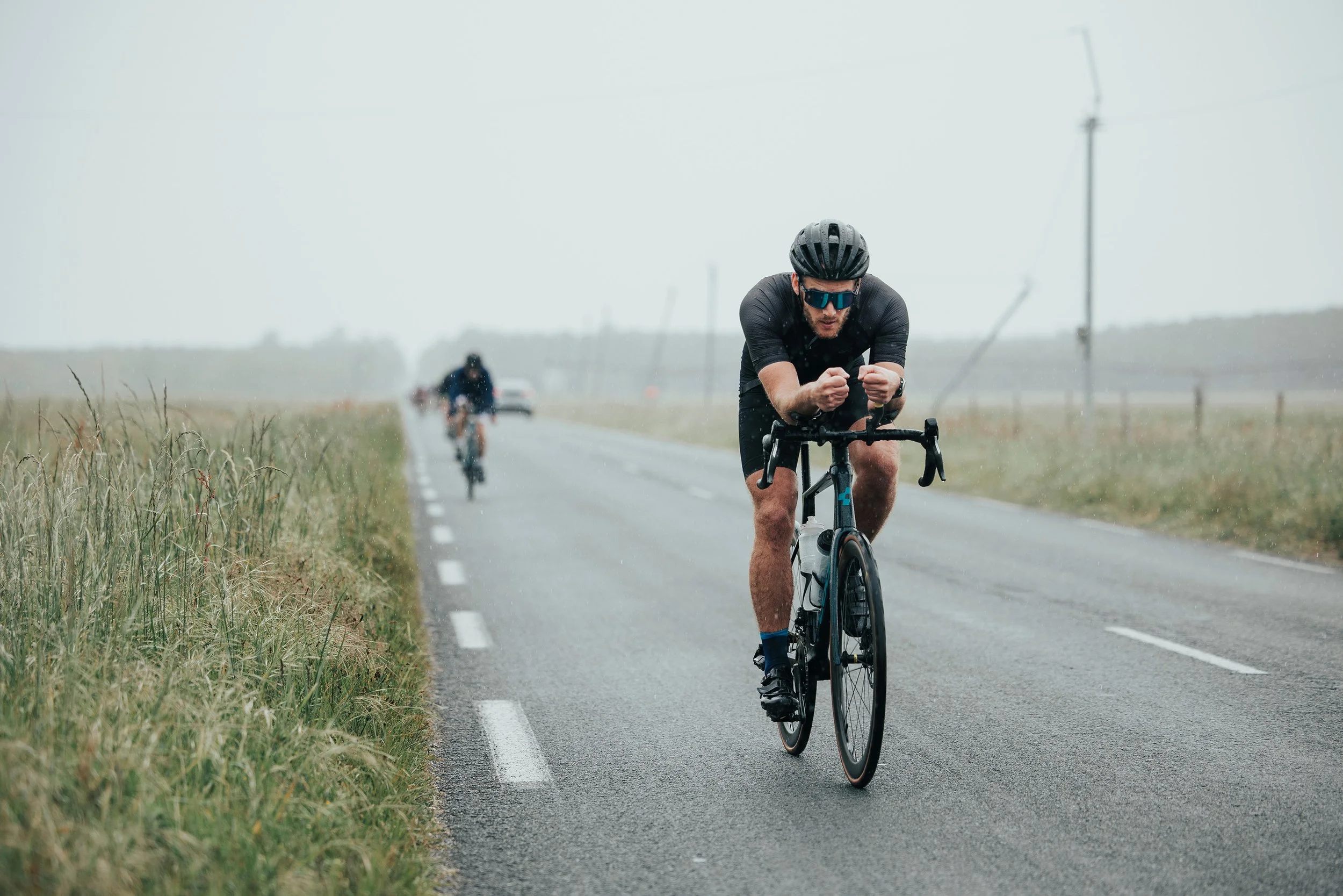 Un cycliste portant un casque et des lunettes modernes, pédalant sous la pluie sur une route rurale avec deux autres cyclistes en arrière-plan.