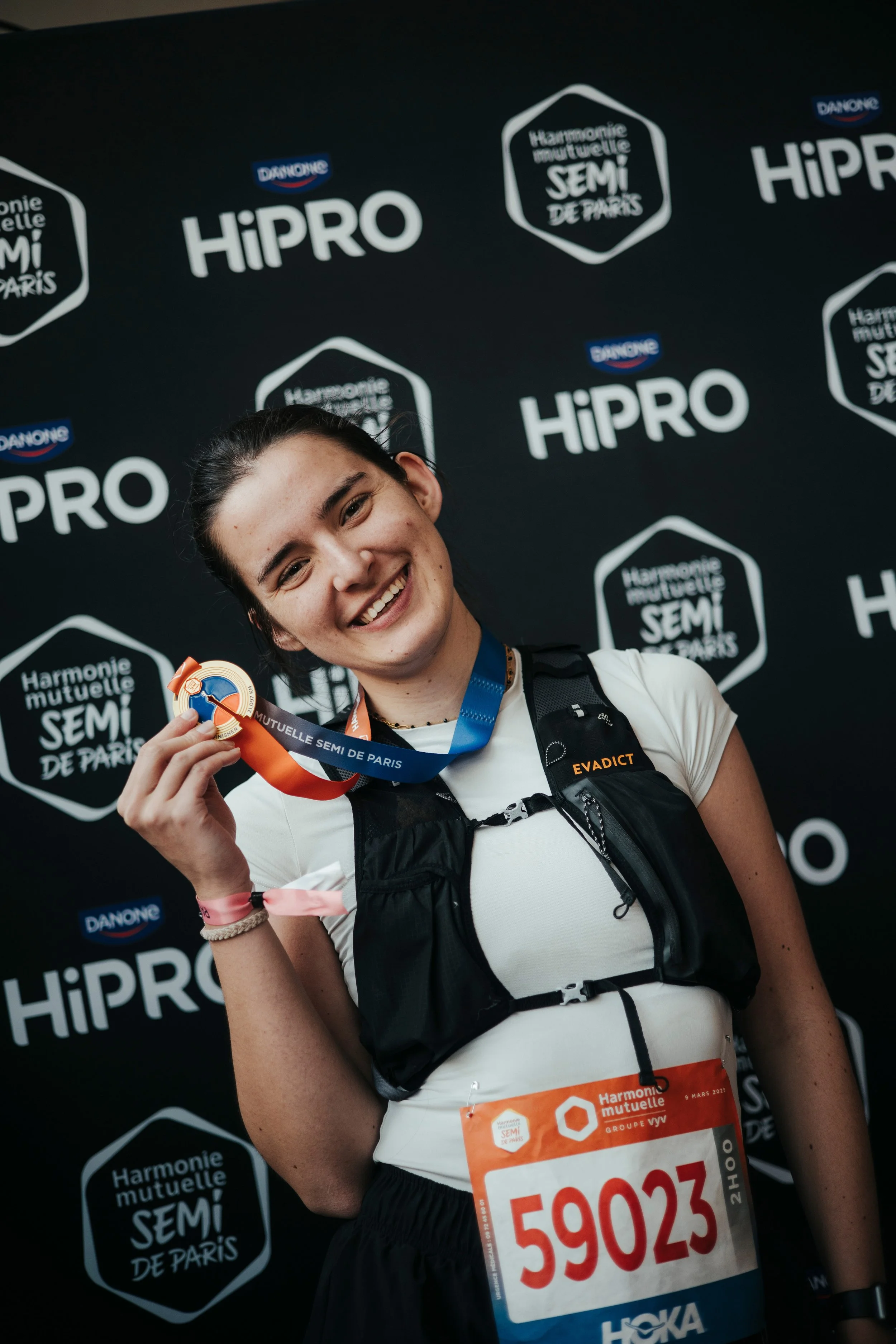 Une jeune femme souriante, portant un dossard et une médaille, après avoir participé à une course ou un événement sportif, avec un arrière-plan affichant les logos de HIPRO, Danone, et de l'Harmonie Mutuelle Semi de Paris.