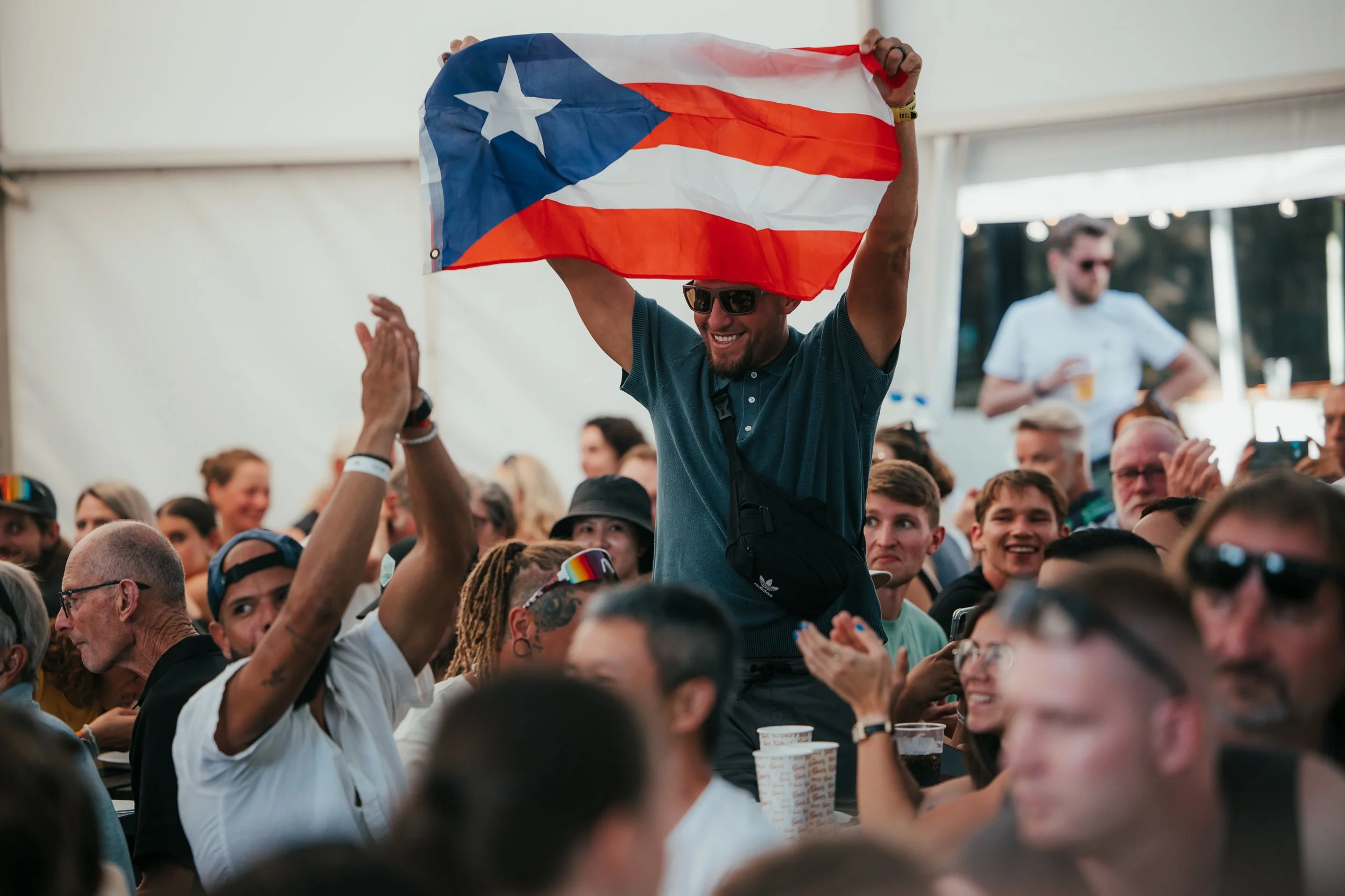 Groupe de personnes assistant à un événement, un homme en haut tient un drapeau de Porto Rico, tout le monde semble heureux et applaudit.