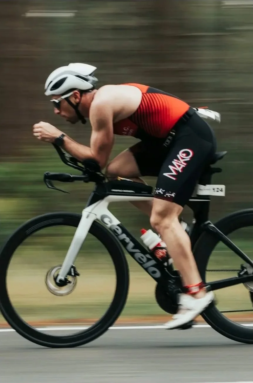 Un homme faisant du vélo de course sur une route, portant un casque blanc, un maillot sans manches rouge et noir, et des shorts noirs avec des logos.