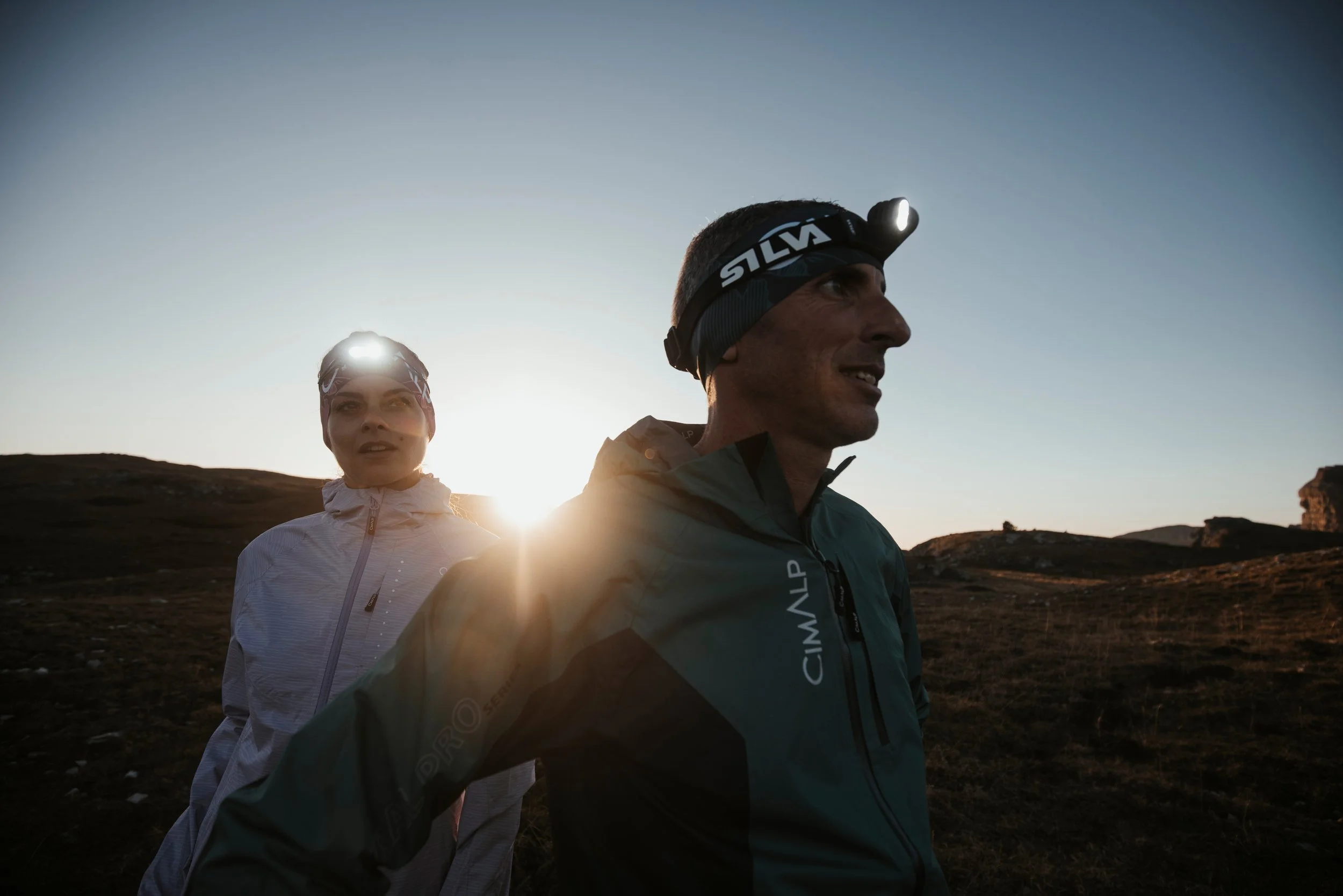 Deux personnes en vêtements de sport, portant des lampes frontales, marchant ou courant dans un paysage de collines au lever ou coucher du soleil.