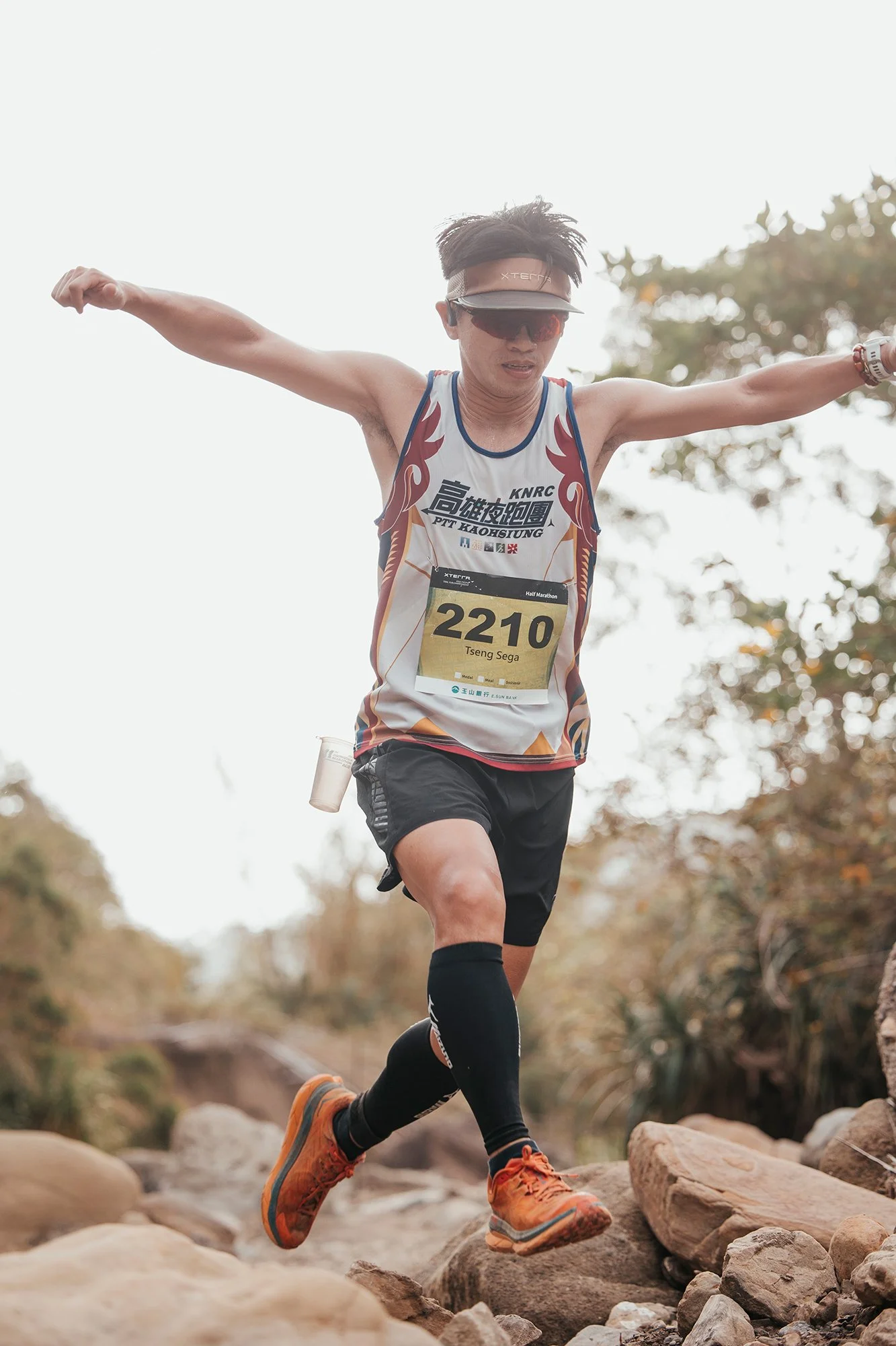 Un homme participe à une course en pleine nature, sautant sur des rochers, portant un t-shirt de course, un casque, des lunettes de soleil, et un numéro de course 2210.