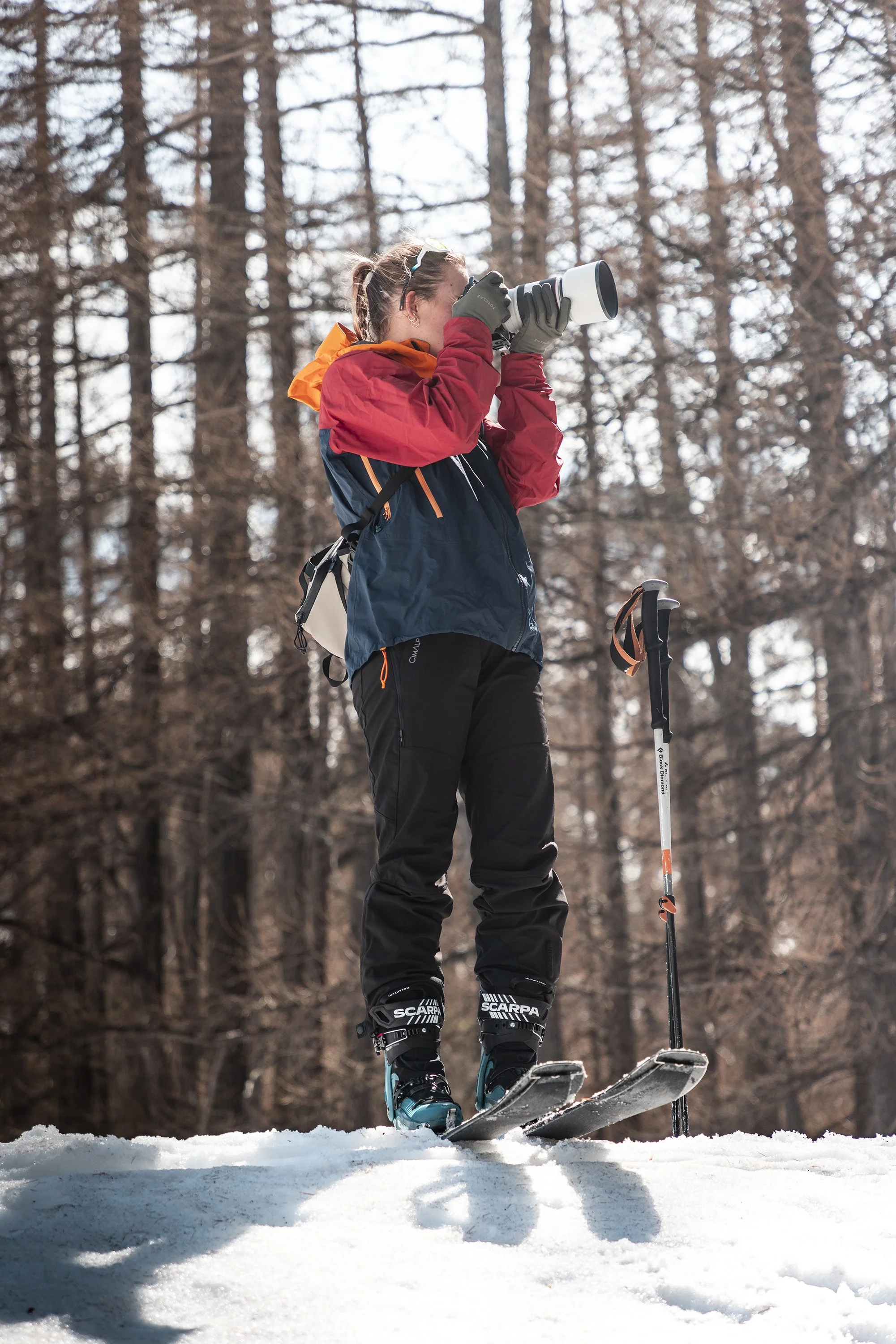 Une femme en vêtements de ski tenant un appareil photo, debout sur la neige devant un forêt d'arbres. Des chaussures de skis SCARPA aux pieds.