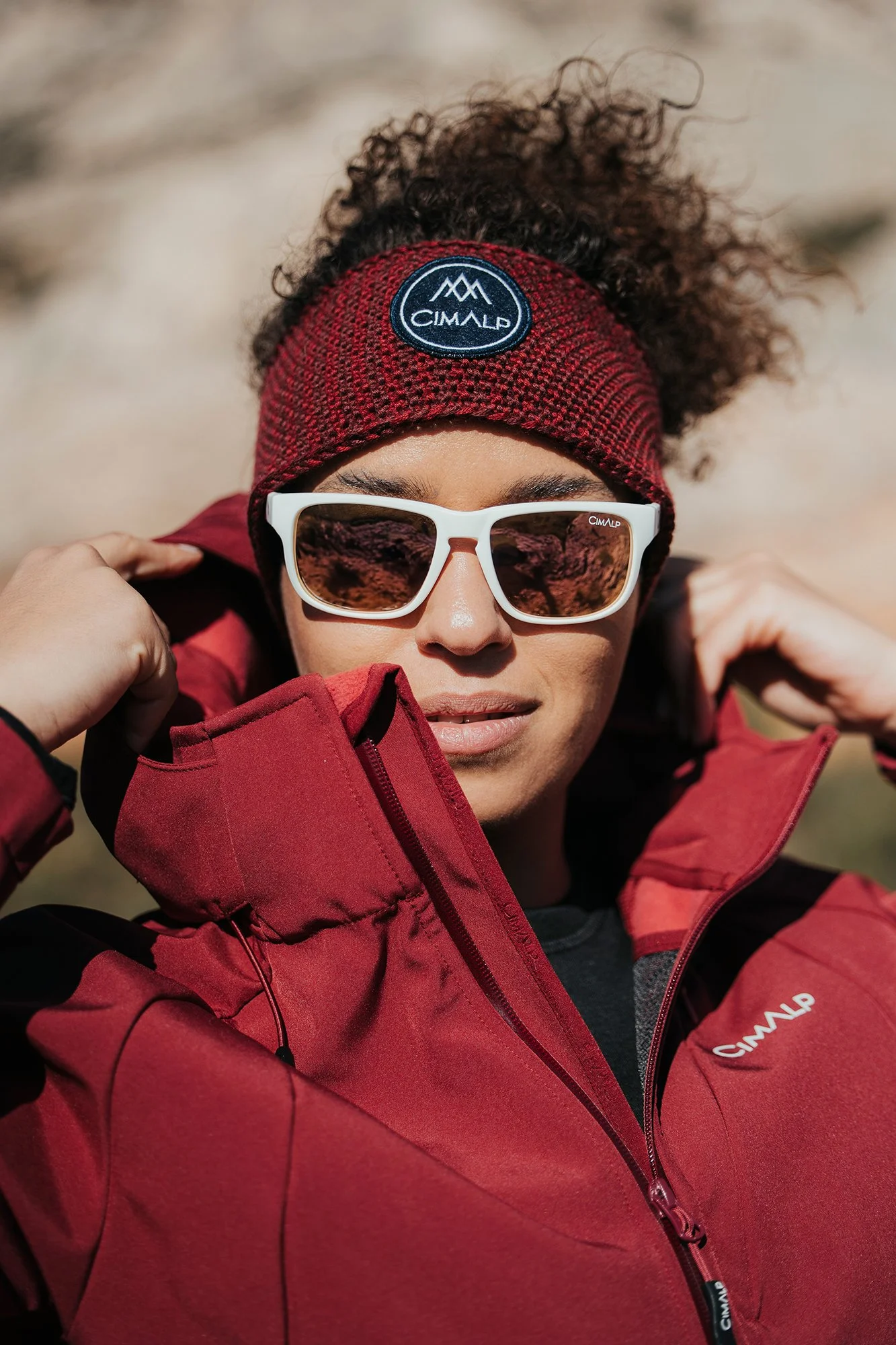 Une femme portant des lunettes de soleil blanches, une veste rouge et un bonnet rouge foncé avec un logo "CIMALP", dans un environnement en plein air.