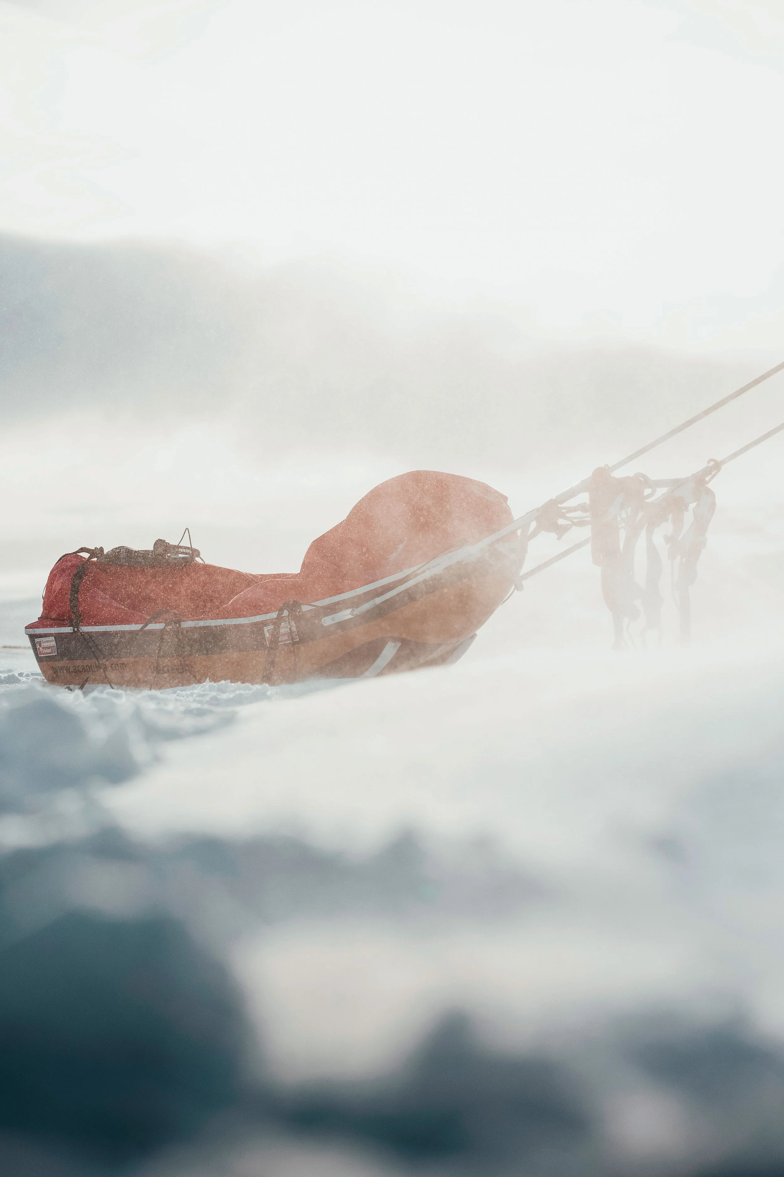 Une embarcation de sauvetage orange sur de la glace, tirée par des personnes vêtues d'équipement d'expédition dans un environnement glacé et venteux