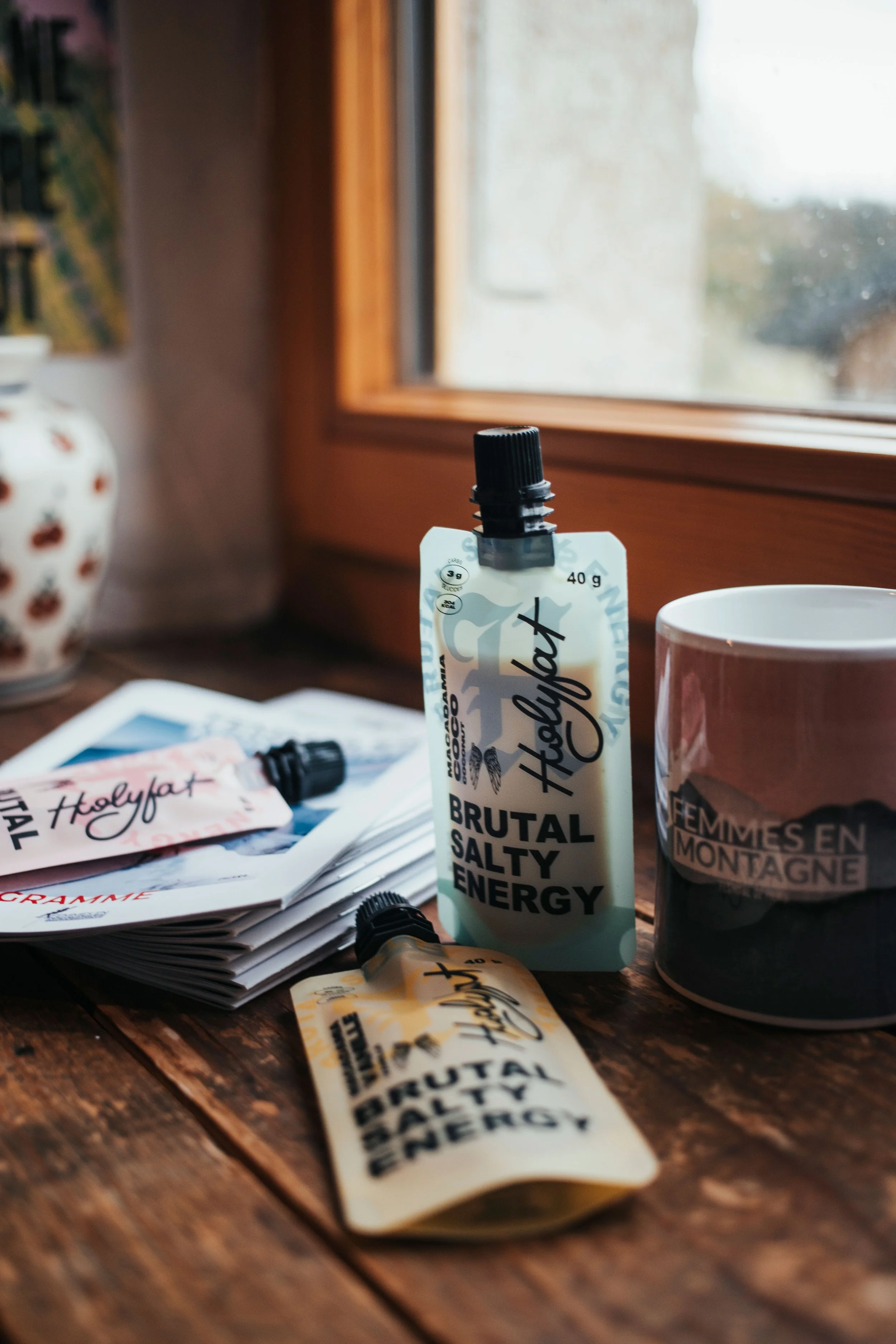 Tubes de crème énergisante Holyfat sur une table en bois, à côté d'une tasse et de papiers, près d'une fenêtre