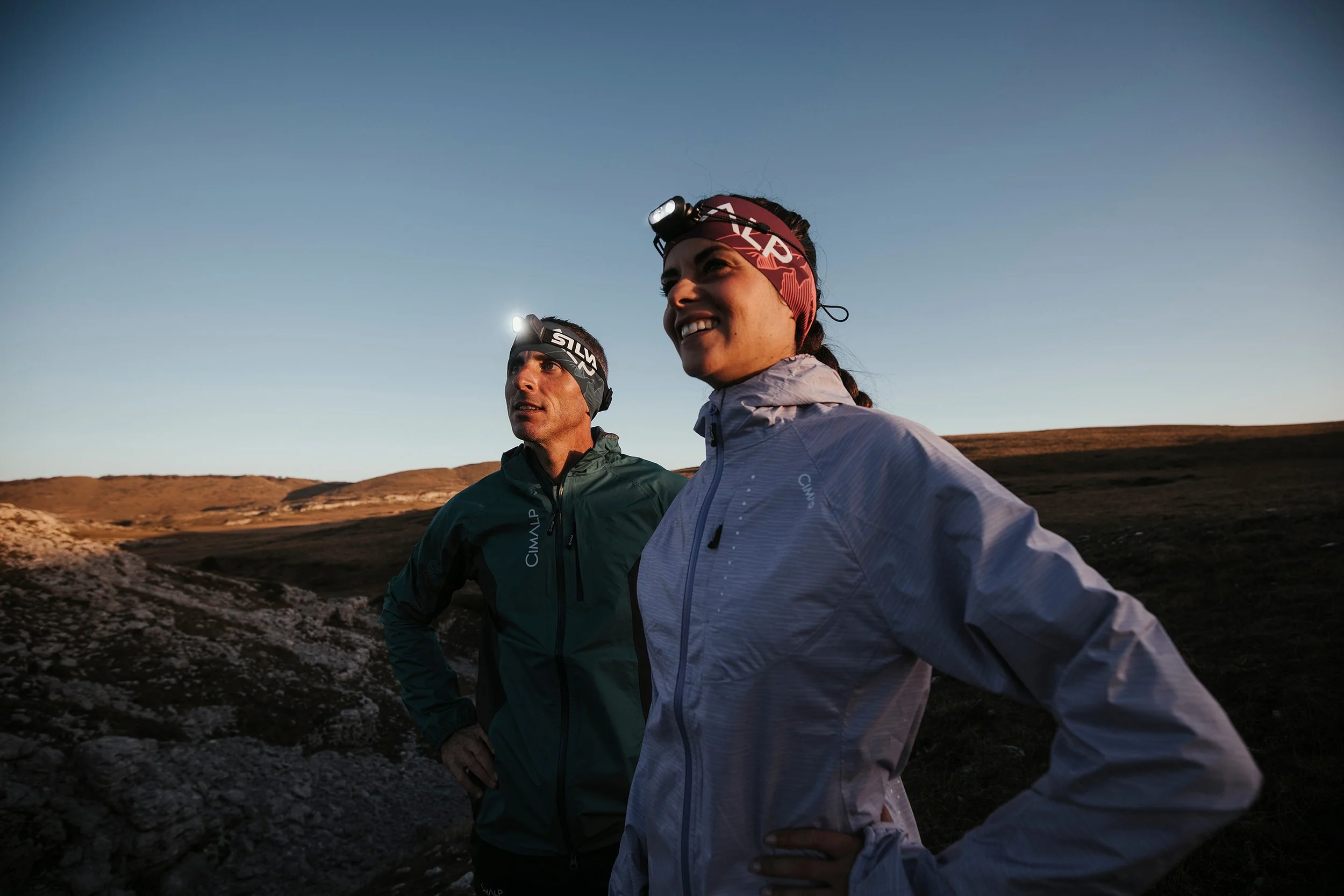 Deux personnes en vêtements de sport avec des lampes frontales, regardant vers l'horizon dans un paysage de collines au crépuscule.