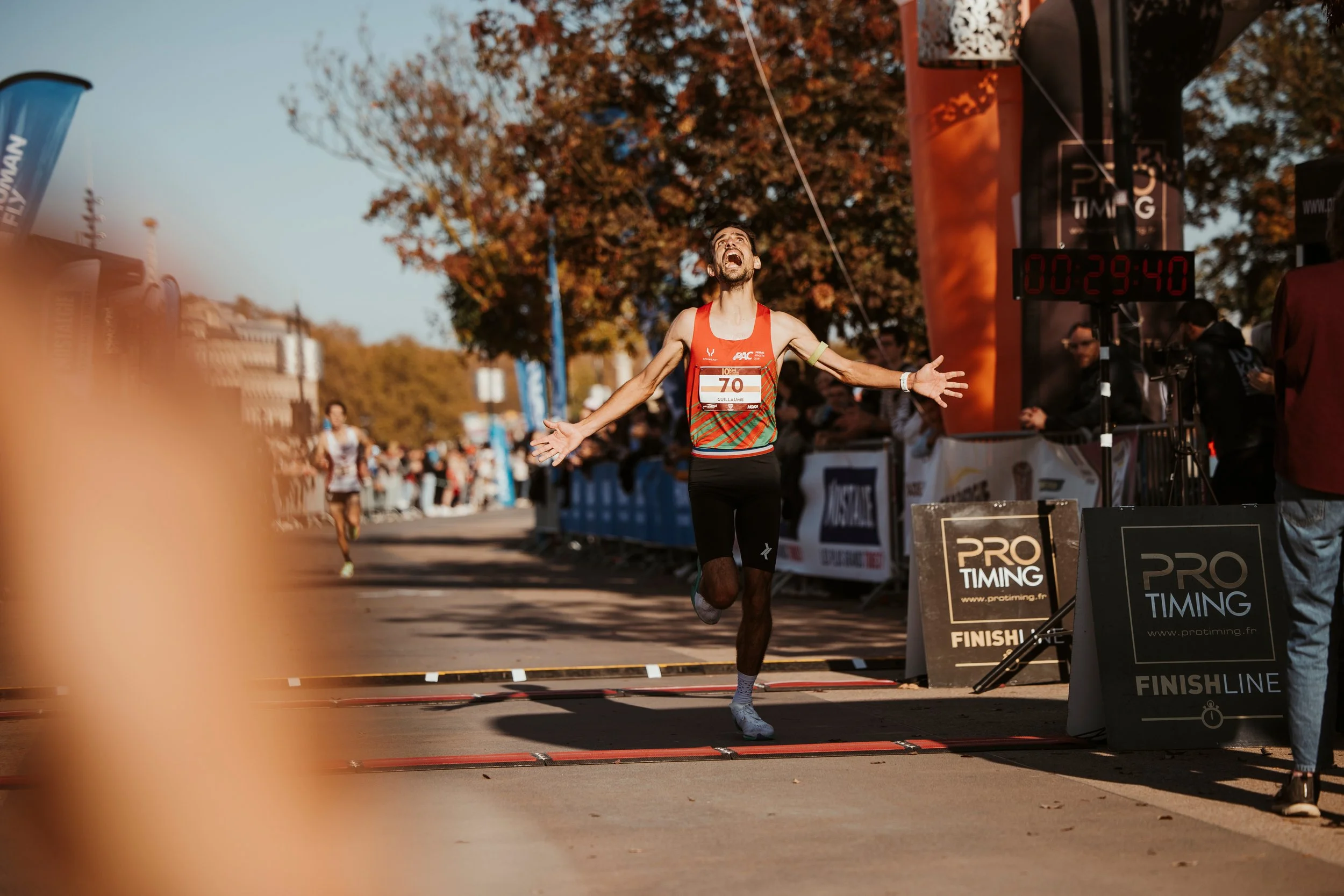 Un coureur féminin franchit la ligne d'arrivée d'une course, avec les bras ouverts et une expression de joie. Il porte un maillot rouge, noir et vert, et un dossard numéro 70. Le chrono affiche 29:40, et l'événement semble se dérouler en automne, ave