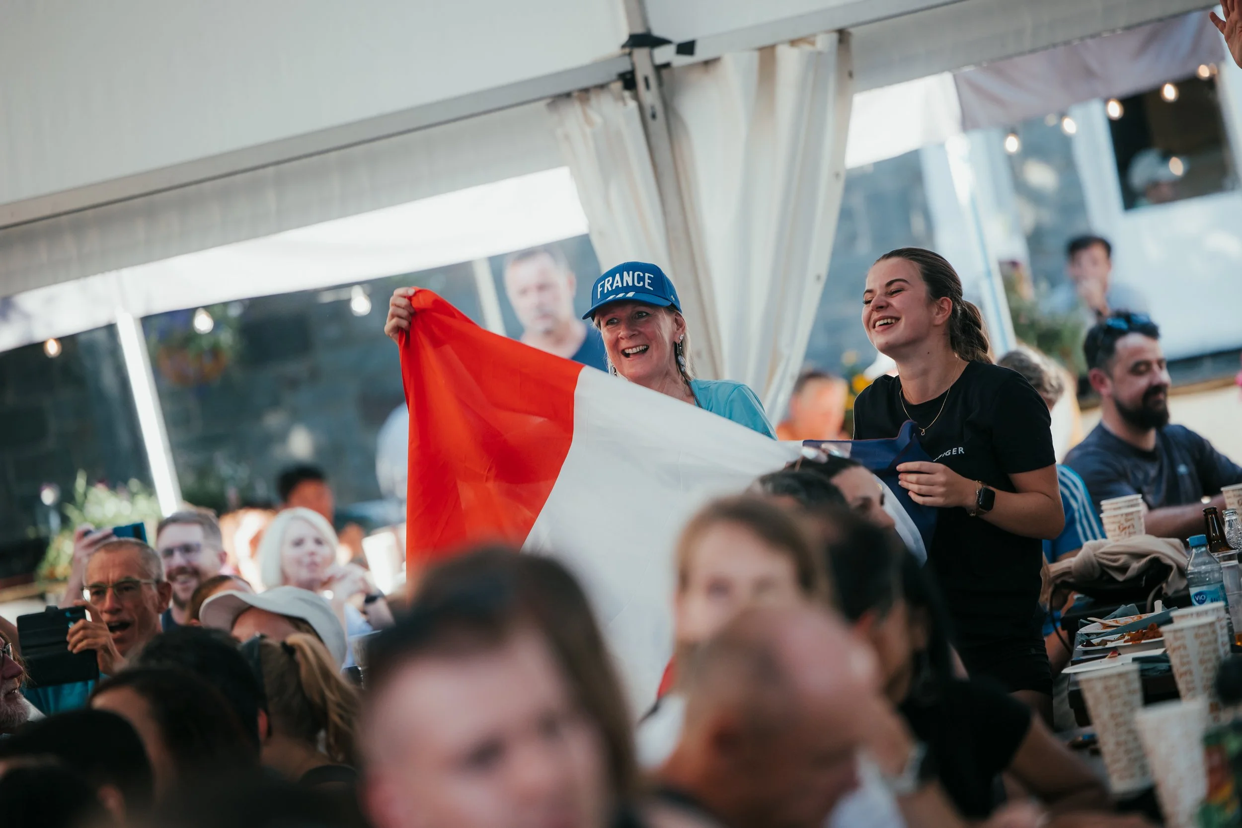 Une femme tient un drapeau français à une fête ou un événement, entourée de plusieurs personnes qui semblent célébrer ou regarder un match.