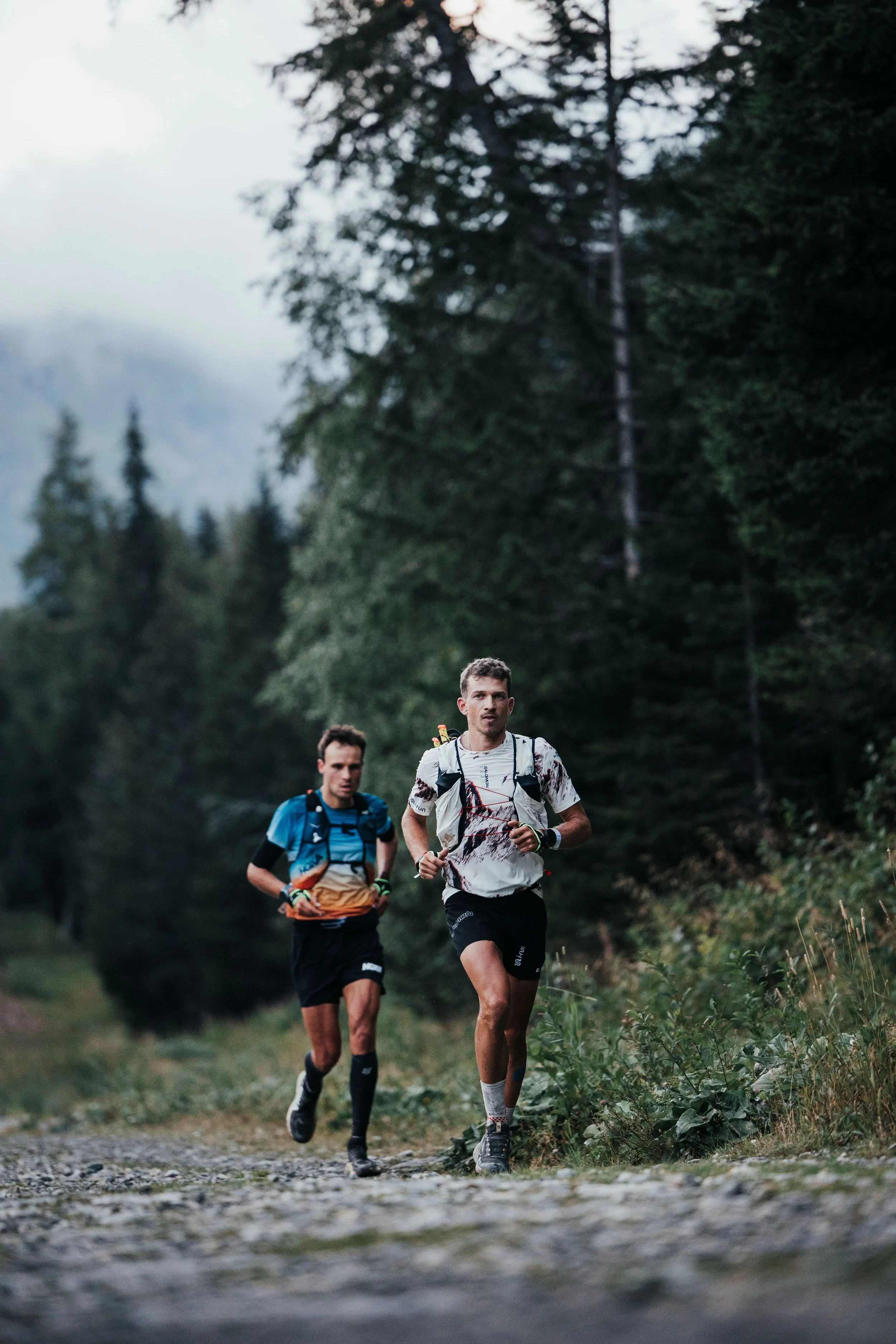 Deux hommes courent sur un sentier en forêt, portant des sacs à dos pour la course ou la randonnée.