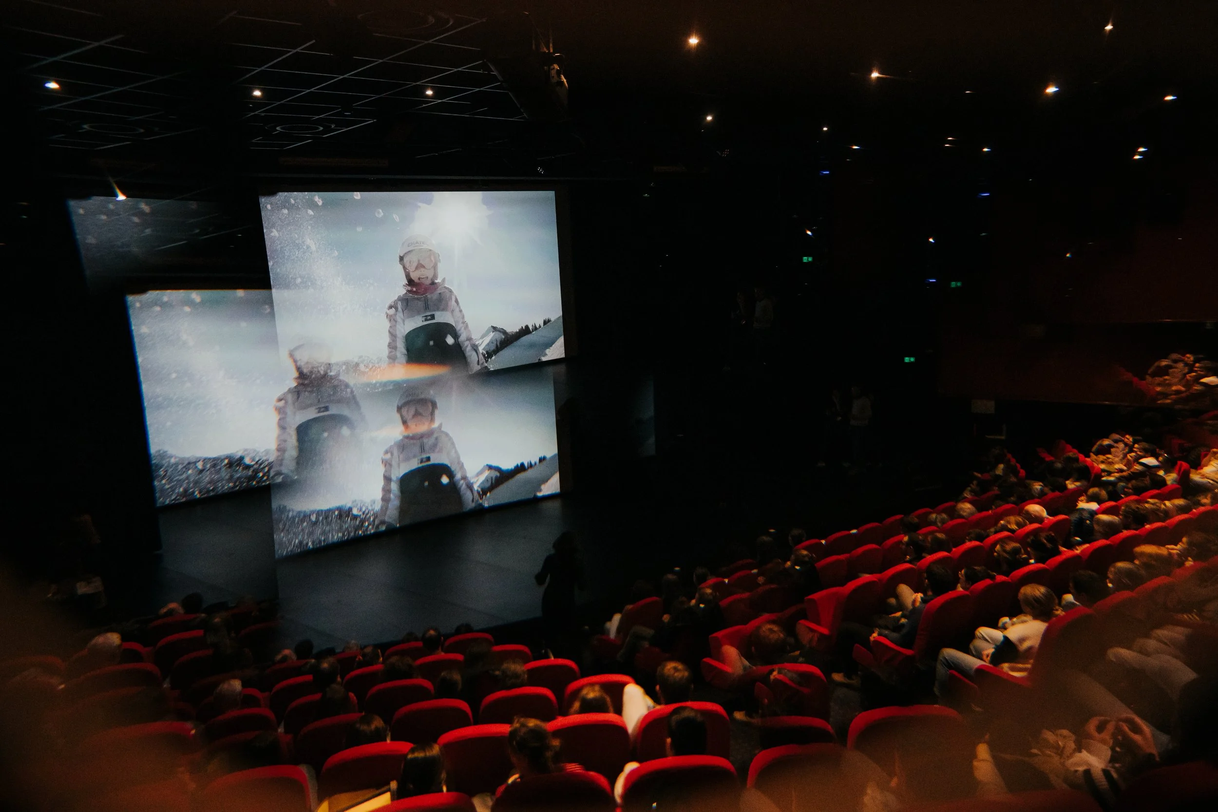Une salle de cinéma remplie de spectateurs regardant un film dans une salle sombre. Deux écrans montrent une scène d'un enfant en tenue de ski avec des lunettes et un casque, avec un paysage enneigé en arrière-plan.