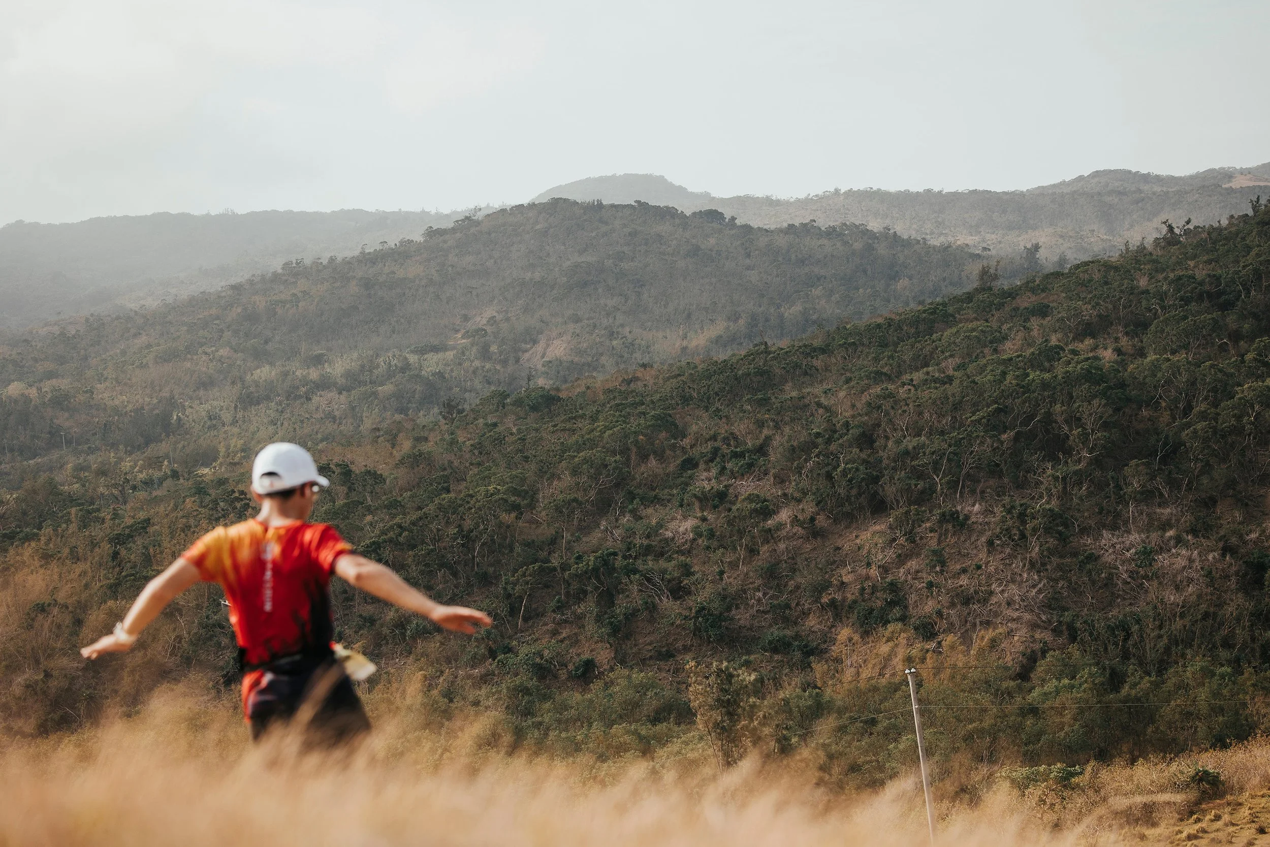 Enfant portant un casque blanc et un t-shirt rouge, courant dans un champ herbeux avec des montagnes en arrière-plan.