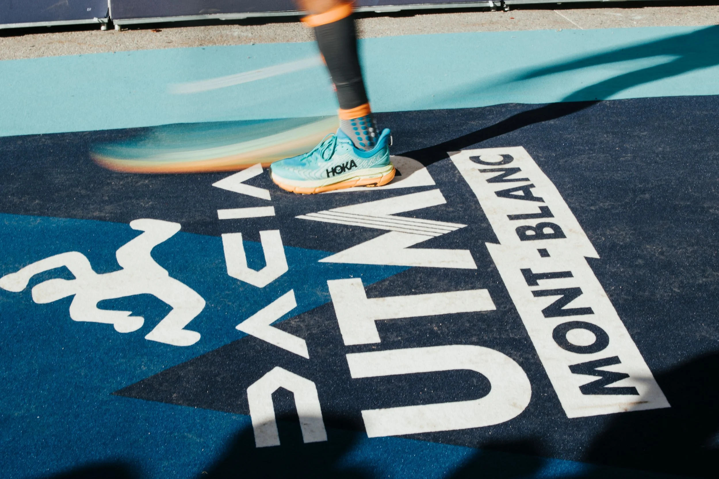 Chaussure de course bleue avec le logo HOKA, en mouvement sur une piste de course avec marquage 'DACIA UTMB MONT BLANC'.