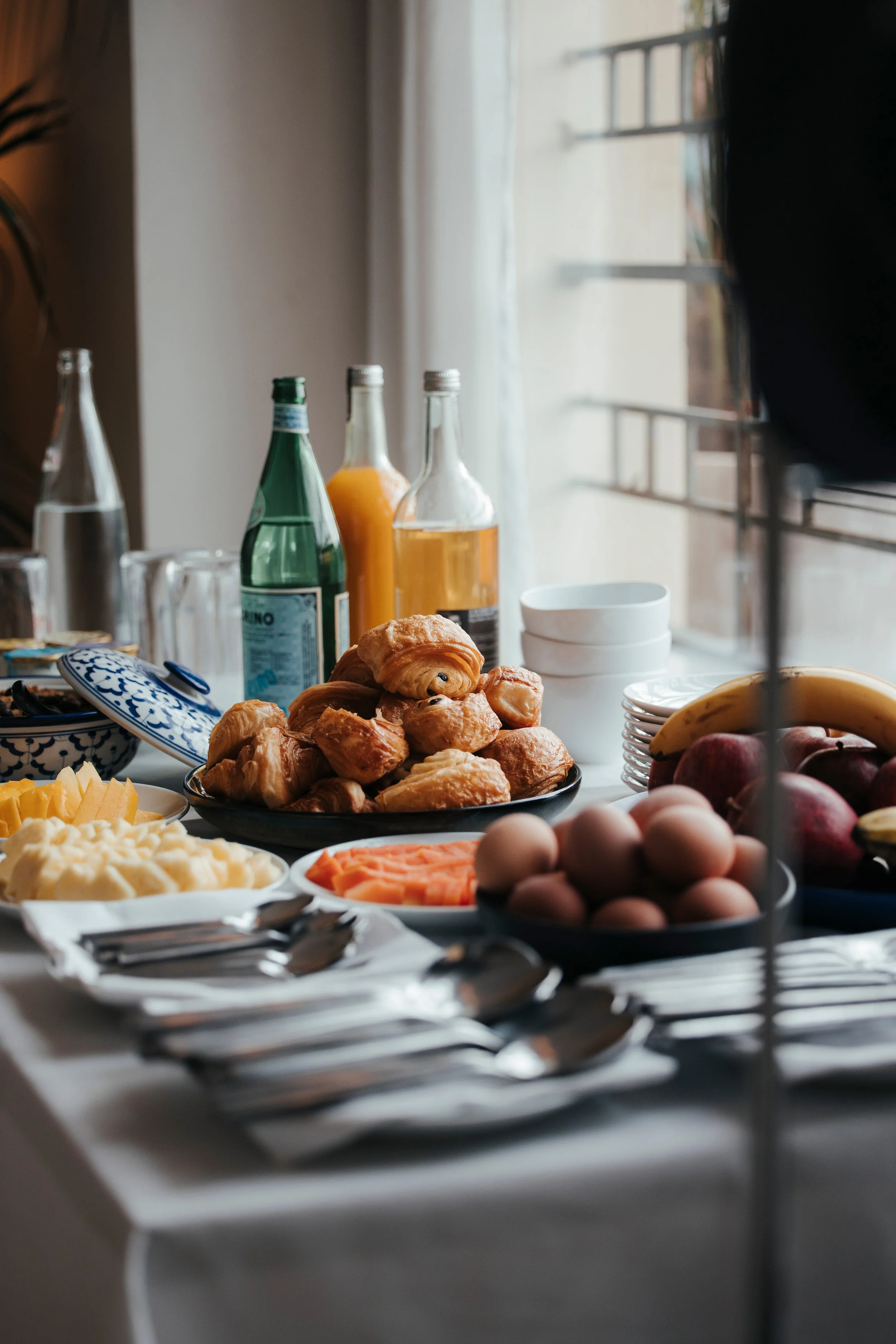 Petit-déjeuner avec croissants, fruits, œufs, jus, et boissons sur une table près d'une fenêtre