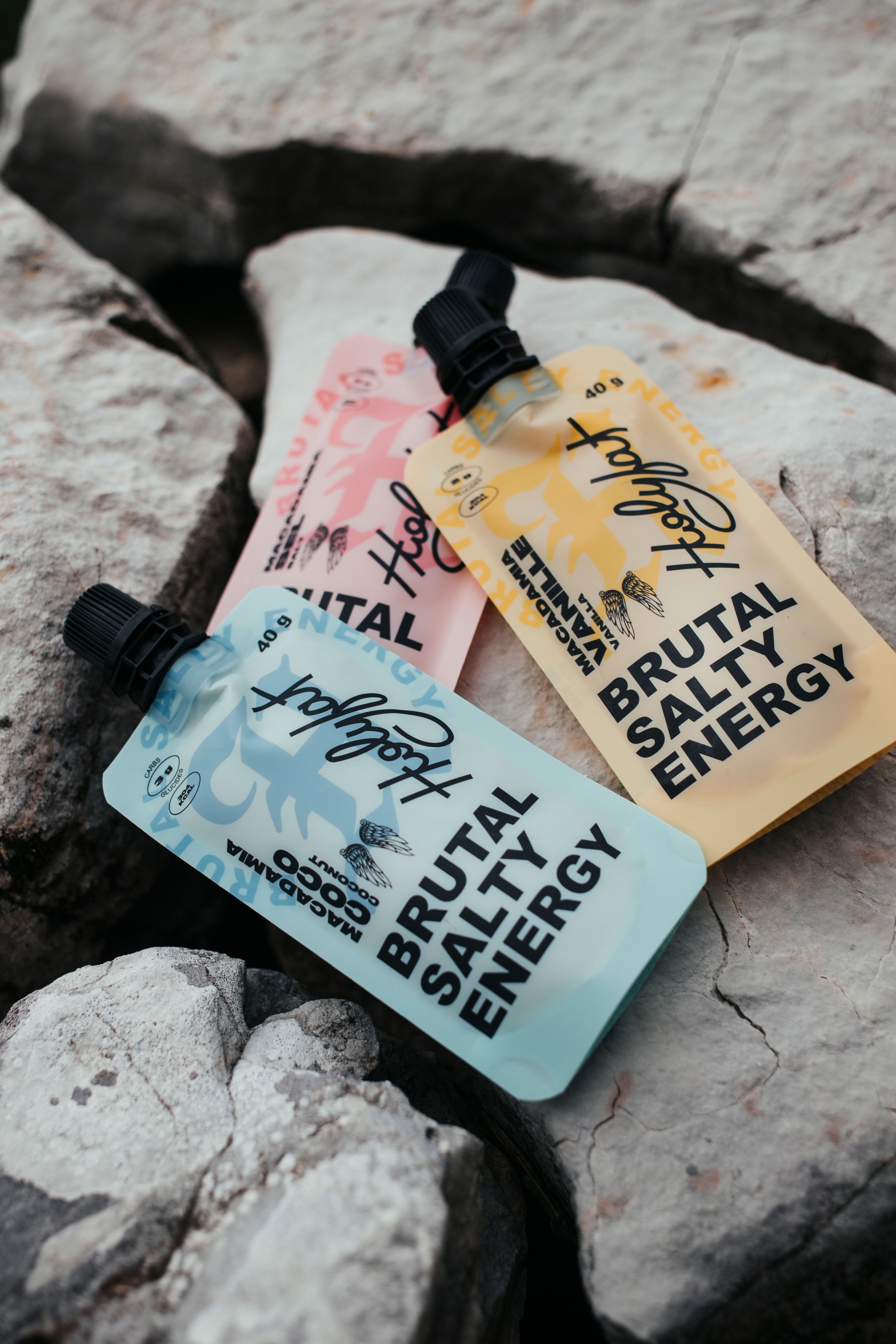 Trois sachets de concentré de boisson énergisante brandée 'Holy Fat' posés sur des rochers. Chacun indique 'Brutal Salty Energy' et contient 40 g, avec des couleurs pastel : bleu, jaune et rose.