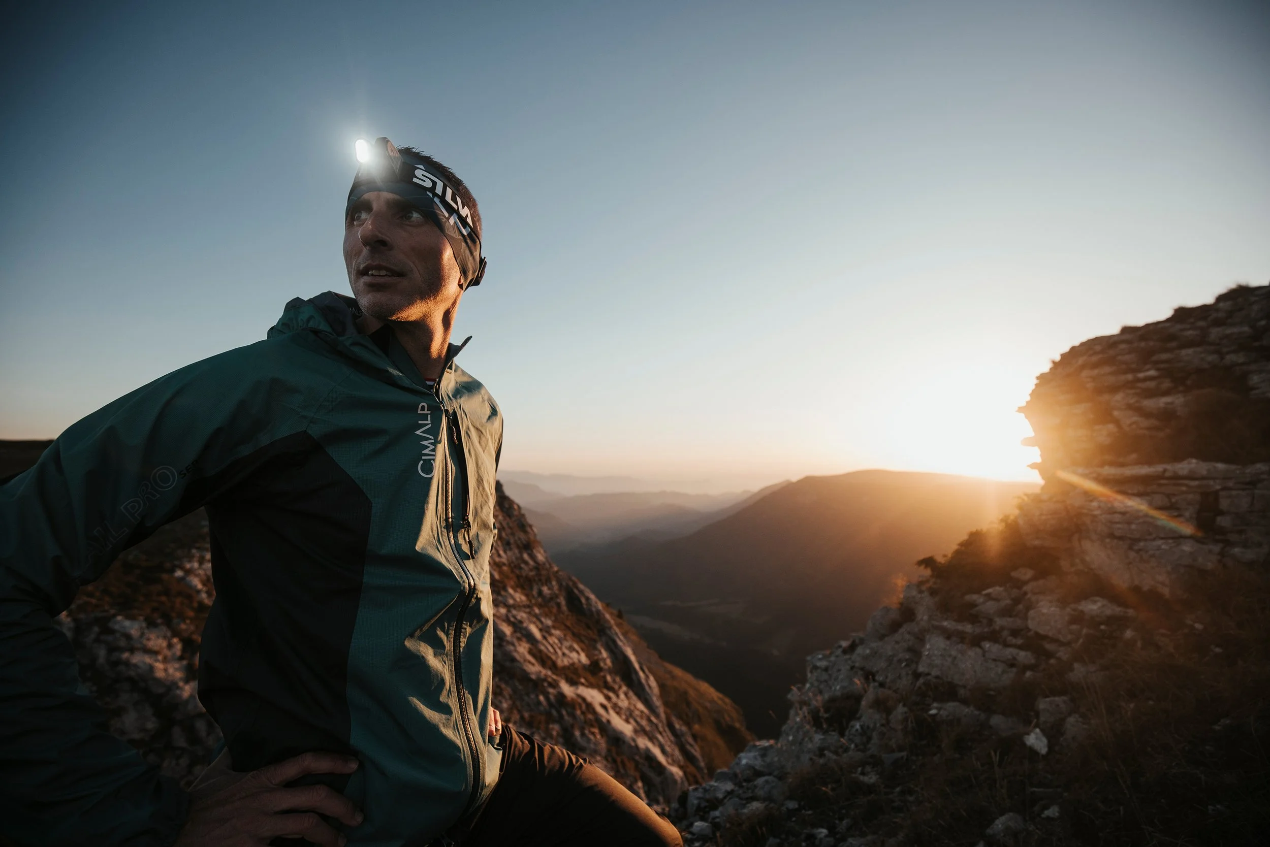 Un homme en pleine nature, portant une veste de sport et une lampe frontale, regarde vers l'horizon lors d'un coucher de soleil dans un paysage montagneux rocheux.