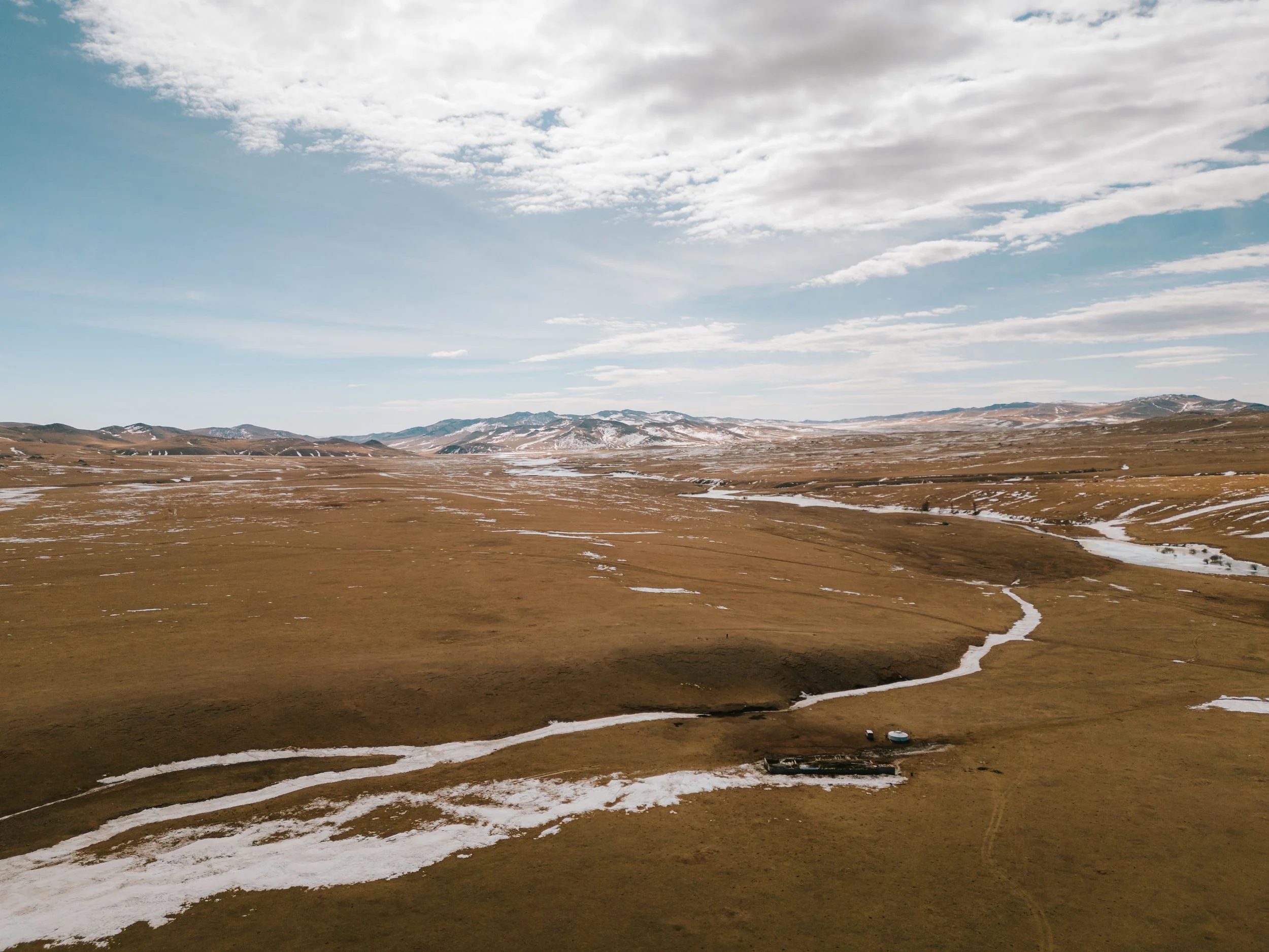 Un paysage de plaines avec des rivières serpentant à travers l'herbe, des montagnes en arrière-plan, et un ciel partiellement nuageux.