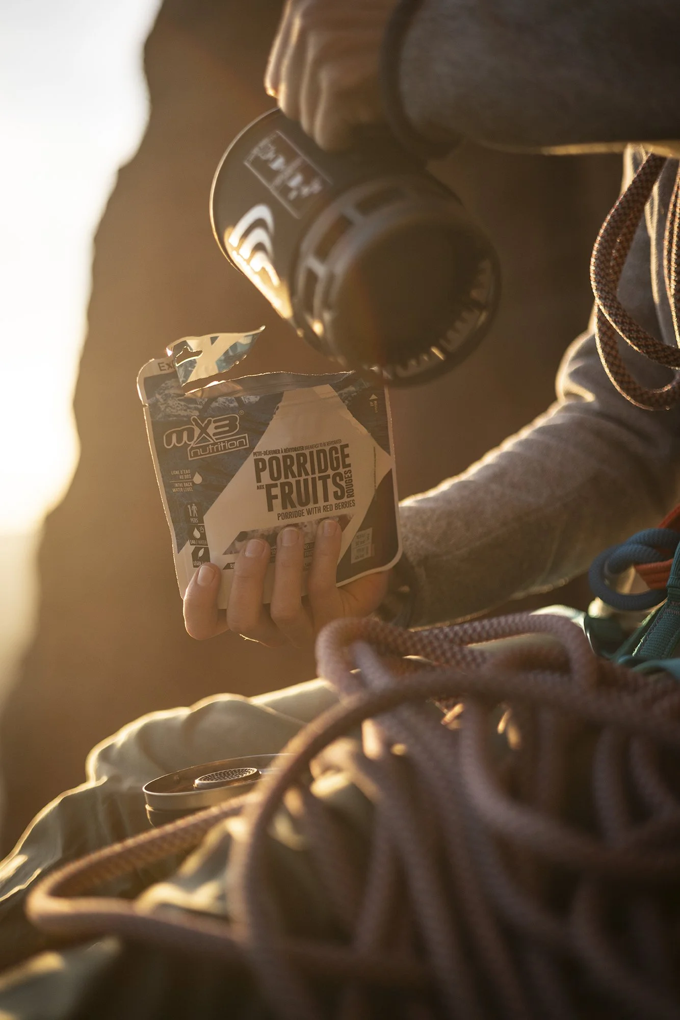 Une personne tenant un bol de porridge aux fruits rouges avec un thermos à proximité, assise en extérieur au coucher du soleil, des chaussures de randonnée visibles en premier plan.