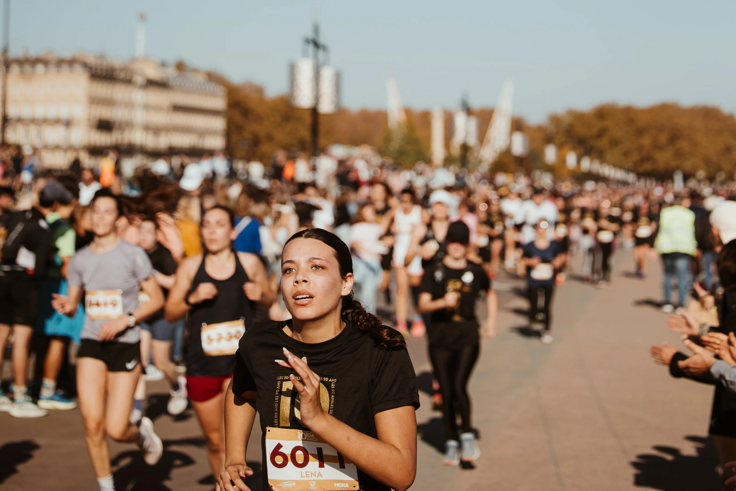 Une femme courante lors d'une course organisée avec de nombreux participants, certains applaudissant, dans une ville avec des bâtiments et des arbres en arrière-plan, sous un ciel dégagé.