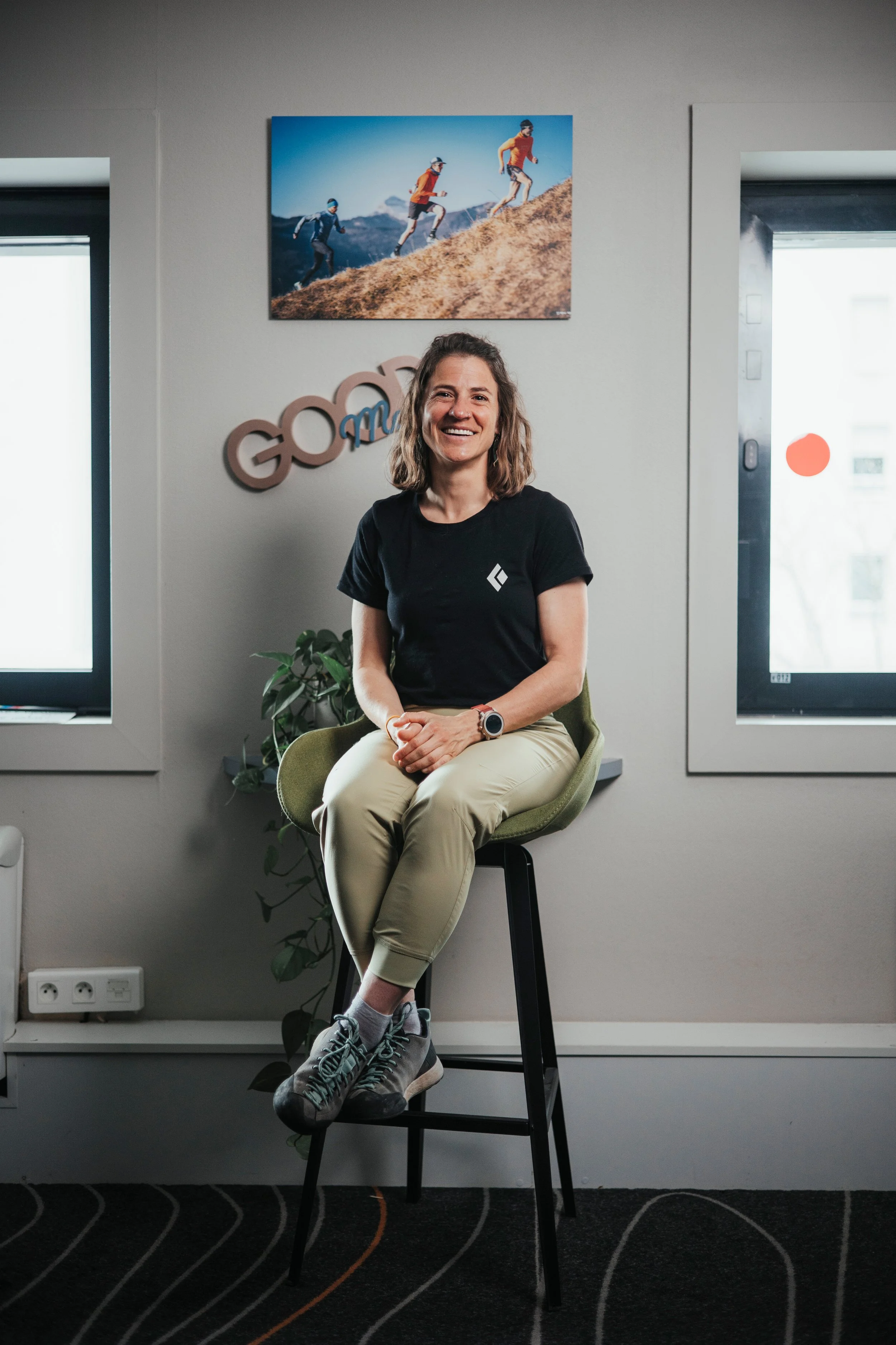 Hillary Gerardi, assise sur un tabouret, souriante, en tenue décontractée dans un bureau moderne avec deux fenêtres, une plante, une photo de marche en montagne sur le mur.